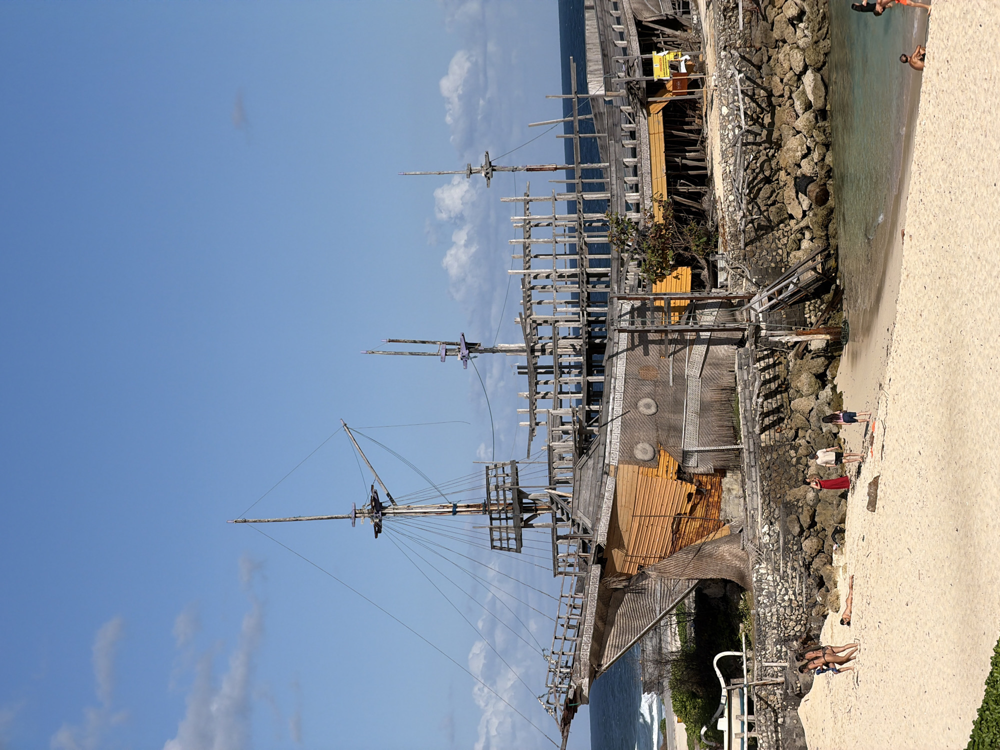 A partially constructed wooden ship structure is situated on the shore in Kuta Selatan, Indonesia. The ship has masts, rigging, and decks made of wood, with some sections covered in orange material. The structure is supported by rocks and wooden beams, and appears to be under construction or renovation. The ocean is visible in the background with gentle waves. In the foreground, a sandy beach is populated by several individuals. Some are walking, others are sunbathing, and one person is lying on their back. The sky is blue with scattered white clouds.