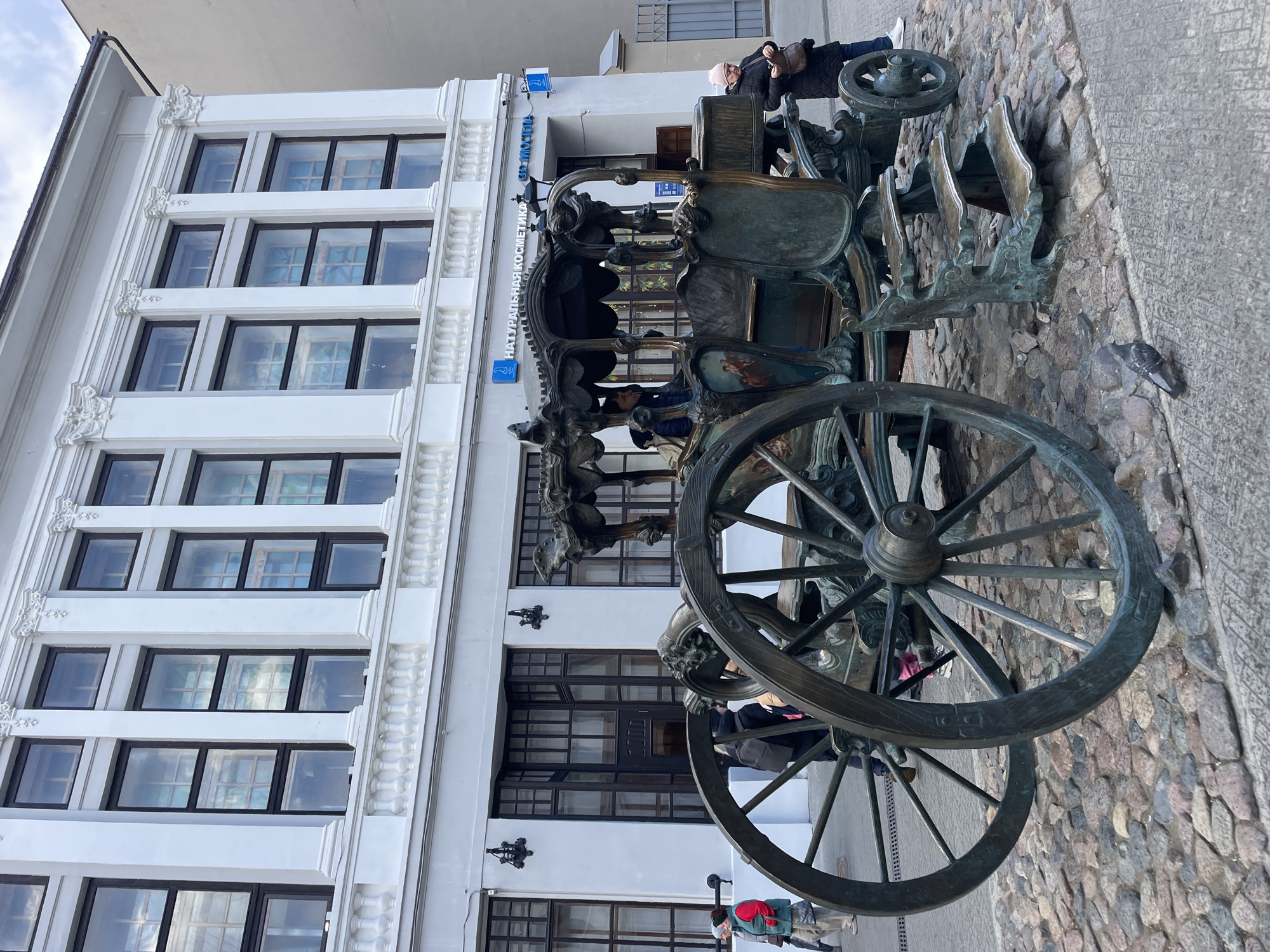 The image captures an urban street scene in Kazan, Russia, featuring a large, ornate bronze sculpture of a carriage or sleigh in the foreground, set against a modern white building. The ground is paved with cobblestones, with a section of patterned concrete to the right.
The central subject is the intricate bronze sculpture, depicting a historical carriage or sleigh. It has large spoked wheels and elaborate decorative elements, including what appears to be figures or designs on the top and sides. One person is visible sitting inside the sculpture, while another person in a black coat and light-colored hat stands near the back of the sculpture on the right. A pigeon is perched on the cobblestones near the sculpture.
The building in the background is a multi-story structure with a clean, white facade. It features numerous large, dark-framed windows arranged in a grid pattern. Decorative architectural elements, including ornate carvings, adorn the vertical and horizontal sections between the windows, particularly visible in the cropped images. Reflections of the sky and potentially opposite buildings are visible in the windows.
Several people are present in the scene. Besides those near the sculpture, another individual wearing a red jacket and a hat is partially visible on the far left, closer to the building's entrance. The scene appears to take place during the daytime under what seems to be clear or partly cloudy skies, based on the reflections in the windows.
Visible text on the building facade, written in Cyrillic, reads "НАТУРАЛЬНАЯ КОСМЕТИКА," which translates to "NATURAL COSMETICS." Below this, partially obscured, is "МОСТ," likely part of a larger word or address. Blue logos or icons are also present next to the text. The overall impression is a bustling urban environment with historical and modern elements.