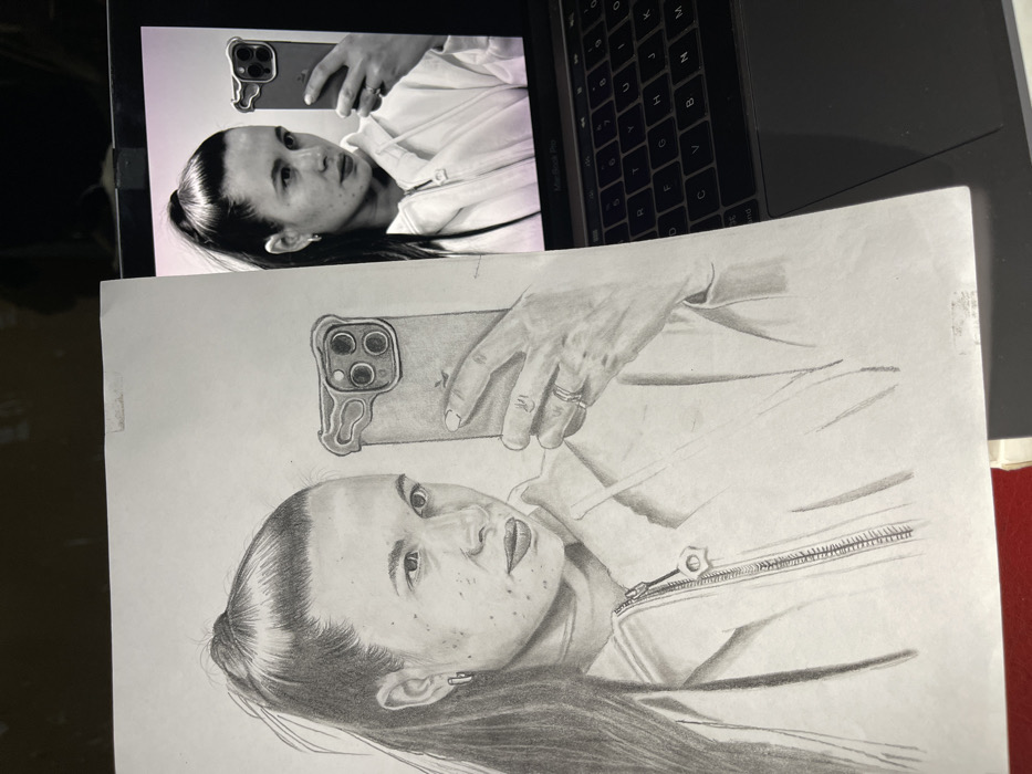 The scene displays a detailed graphite pencil drawing on white paper, placed over a partially visible laptop. The drawing is a realistic portrait of a light-skinned woman with dark hair tied in a high ponytail, wearing a zip-up hoodie and small hoop earrings. She is depicted holding an Apple iPhone with a triple-lens camera and an irregularly shaped clear case, as if capturing a mirror selfie. Two rings are visible on her left ring finger. The laptop, identifiable as a MacBook Pro by its keyboard, displays a color photograph that serves as the reference image for the drawing, showing the identical woman, pose, attire, and phone against a soft, light-colored background. The drawing paper shows minor wear and smudging. A dark, indistinct background surrounds the laptop and drawing, with a hint of a red object beneath the bottom left of the paper. There are no discernible visual elements within the image to forensically confirm its location in the city of Jos, Nigeria.