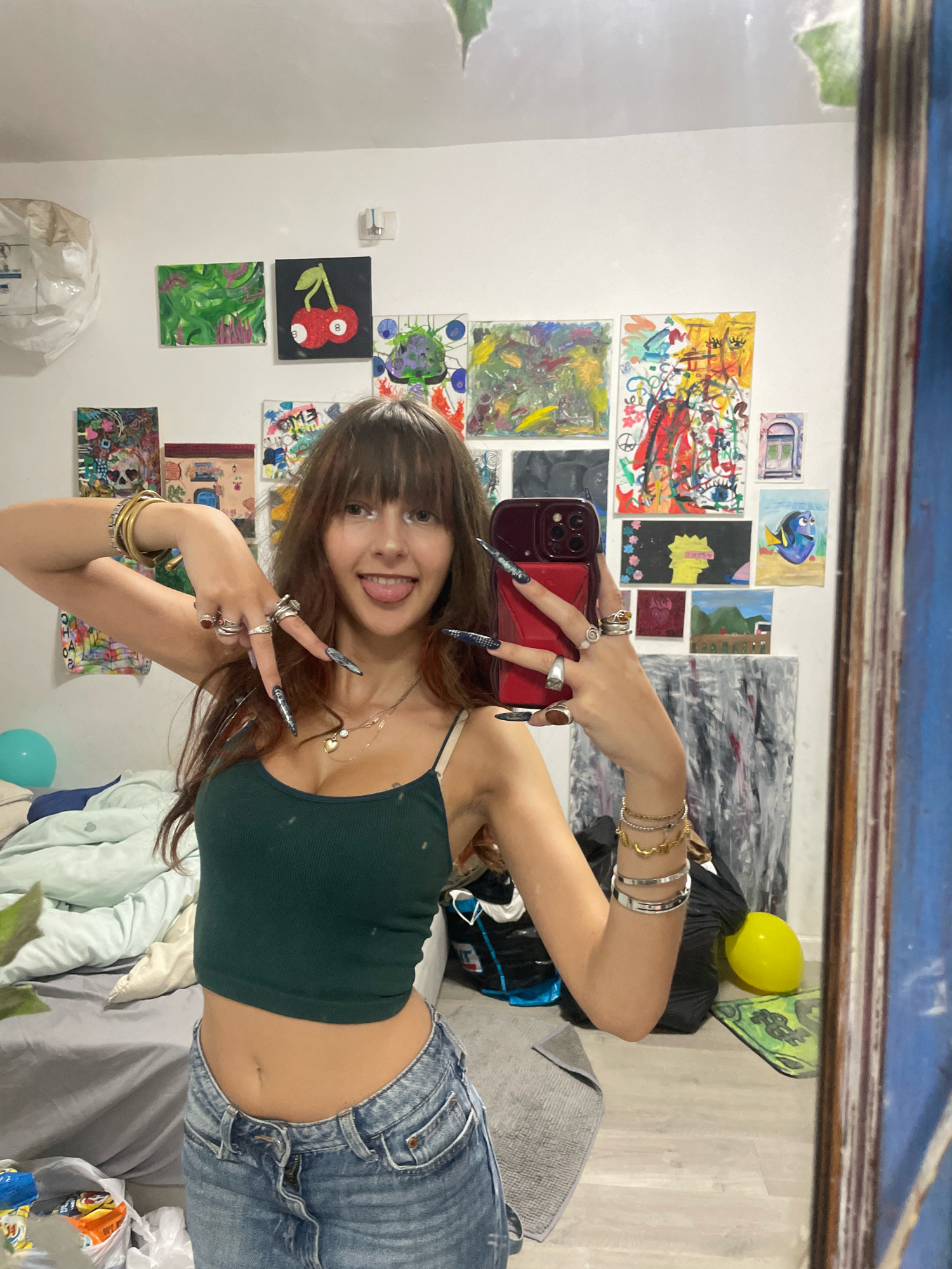In a cluttered bedroom, a young woman with long, brown hair and bangs takes a selfie in a mirror. She wears a dark green crop top and distressed denim jeans. Her right hand, adorned with long, sharp nails and multiple rings, holds a red phone. Her left hand, also decorated with long nails and rings, is raised in a peace sign. She sticks her tongue out playfully at the camera. The room's walls are covered with various colorful paintings and artwork. A bed with rumpled sheets is visible in the background, along with a blue balloon. The overall scene suggests a casual, artistic, and somewhat chaotic personal space. The image does not provide any information that would indicate the location being Raanana, Israel.