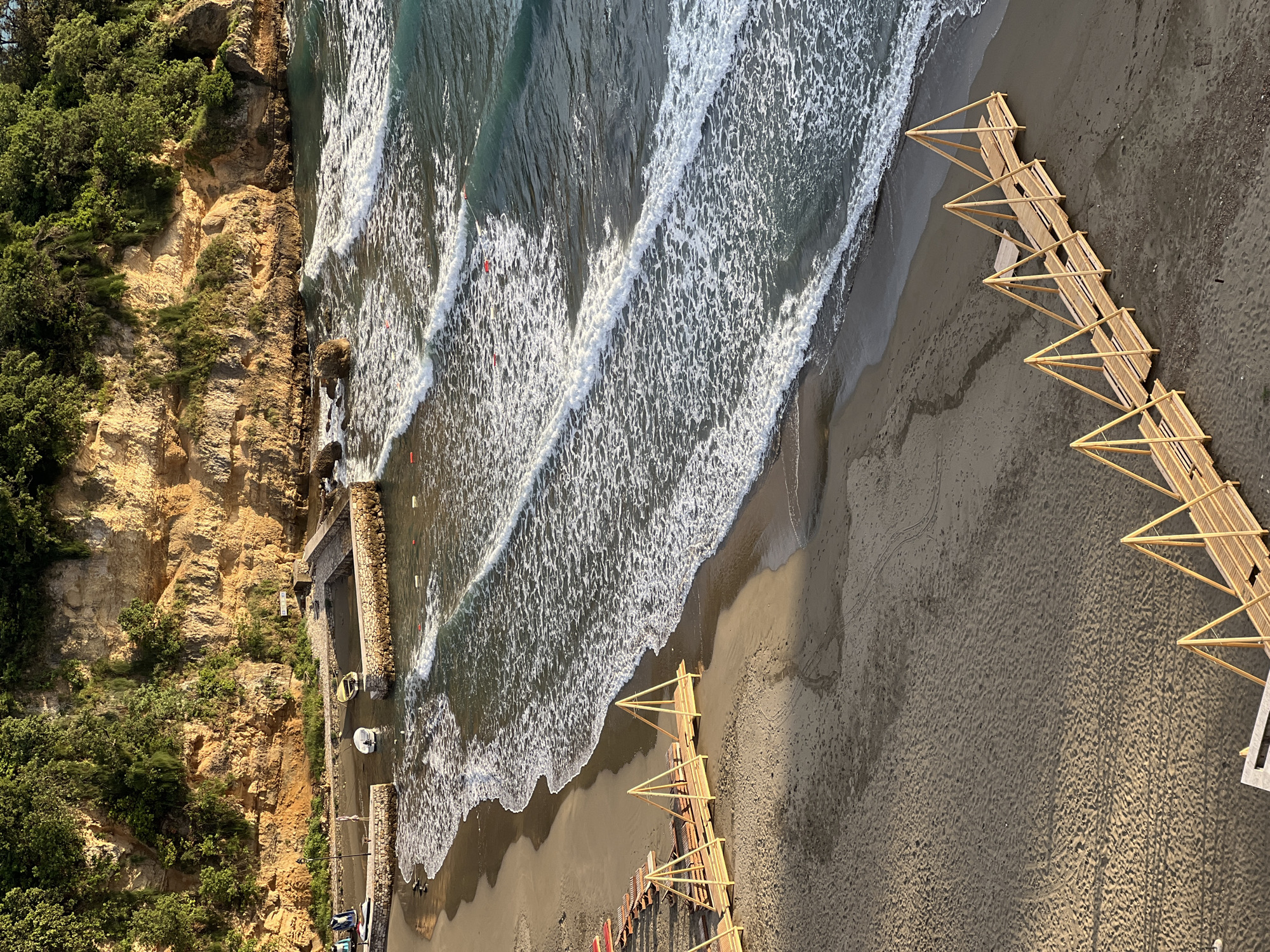 The photograph presents a bird's-eye view of a beach in Ulcinj - Ulqin, Montenegro, featuring a cliff, the sea, and a beach structure. The left side of the image showcases a cliff face covered with vegetation. A section of the cliff transitions into a walled embankment leading into the water. The sea is agitated with visible waves. A distinctive, lengthy, wooden beach structure is set on the sand, extending from the upper right towards the lower center of the frame.