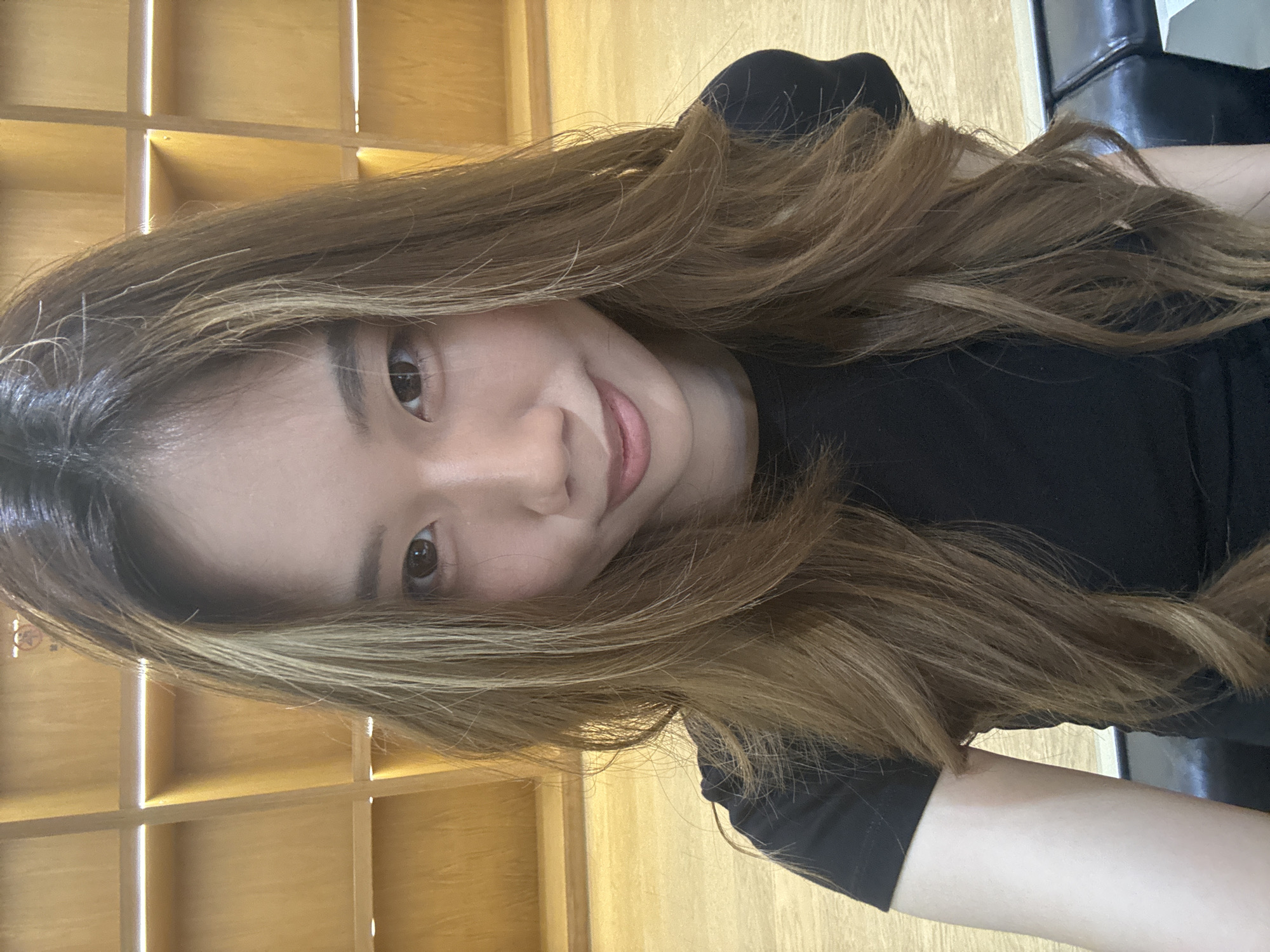 A young Asian woman with light brown hair is centered in the image, smiling subtly at the camera. She's wearing a black top. The background features a grid-like wooden structure, possibly a shelving unit. The setting appears to be indoors. There is no verifiable information in the image connecting it to the city of Nan'an District, China.