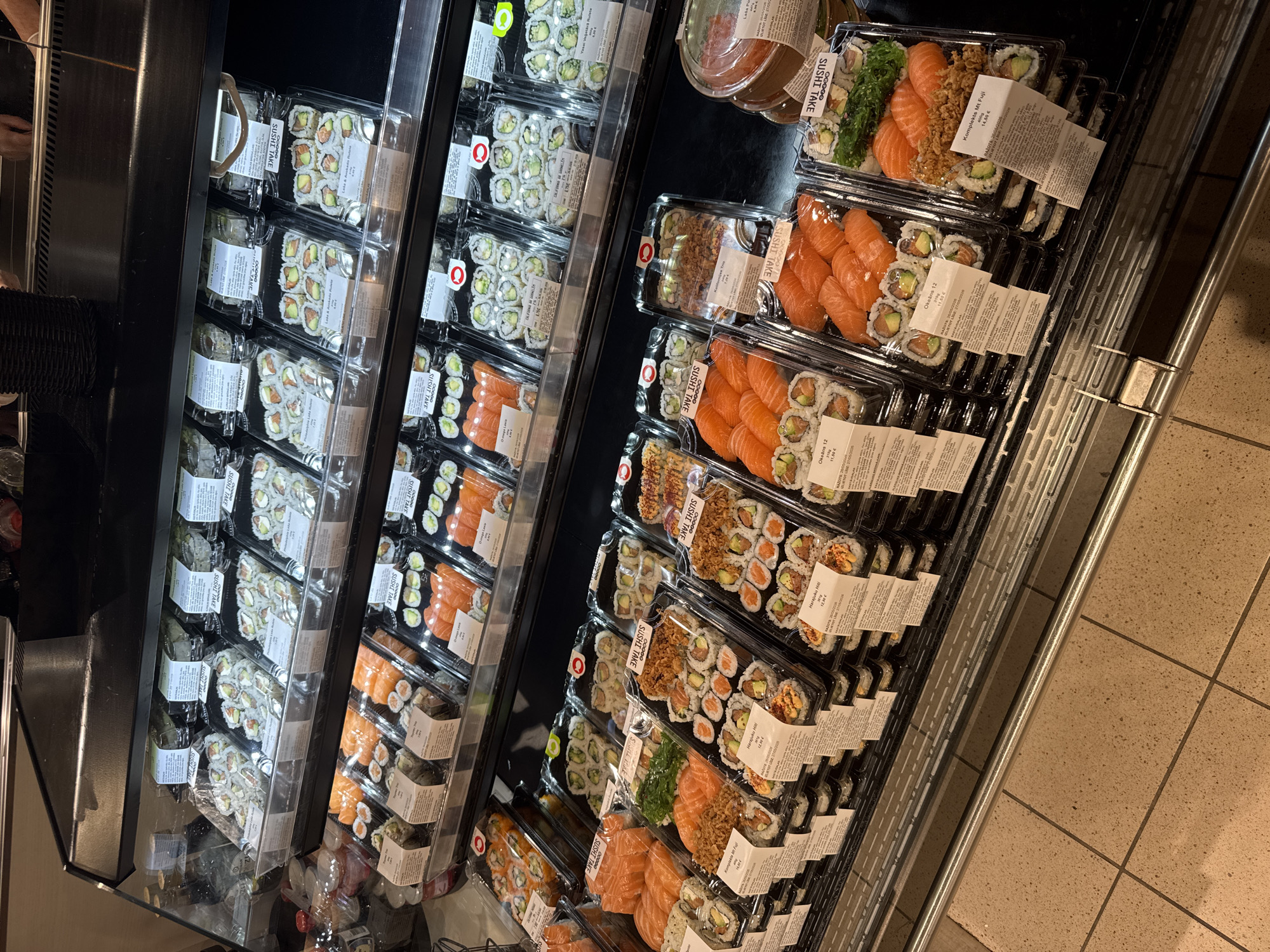 The image shows a refrigerated display case filled with pre-packaged sushi. The display case appears to be located in a store, possibly a grocery store or a supermarket. Several packs of sushi are visible with labels including "Okeans 12", "Komplekts Mt Fuji", and "Harajuku Hiti" along with the price in euros. The brand is "SUSHI TAKE". Based on the text in the labels, it's likely the location is Riga, Latvia. The sushi is arranged in stacked plastic containers.