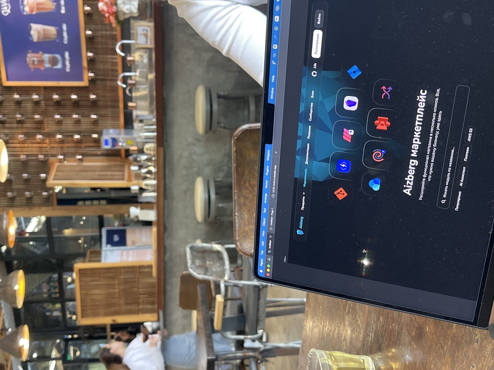 The image shows a person working on a laptop in a cafe. The cafe appears to be in Da Lat, Vietnam based on the Vietnamese text on the laptop screen. The laptop displays a webpage with the title "Aizberg маркетплейс," indicating it's a marketplace or app store. The person is wearing a white shirt, and the background features the interior of the cafe with a wooden bar, stools, and lighting fixtures.