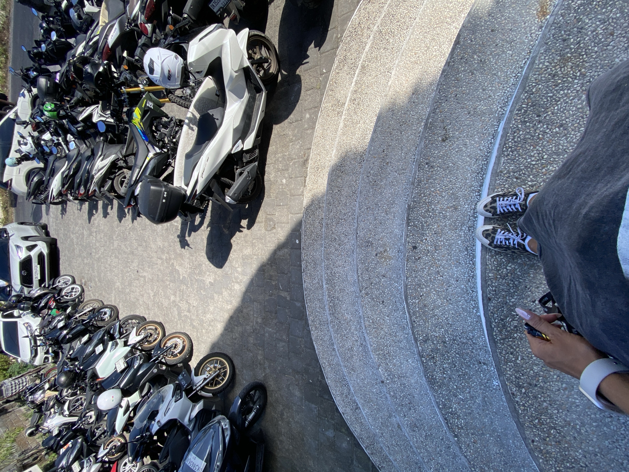 A person stands on a curved concrete stairway in Kuta Selatan, Indonesia, looking down at their feet which are clad in leopard-print sneakers and dark pants. Their right hand, holding a yellow and black object, is visible near the stairs. The stairway overlooks a vast parking lot filled with numerous scooters and motorcycles, some of which are white, black, and silver. In the background, two white cars are parked. The scene is illuminated by bright sunlight, casting shadows across the parking lot and stairs.