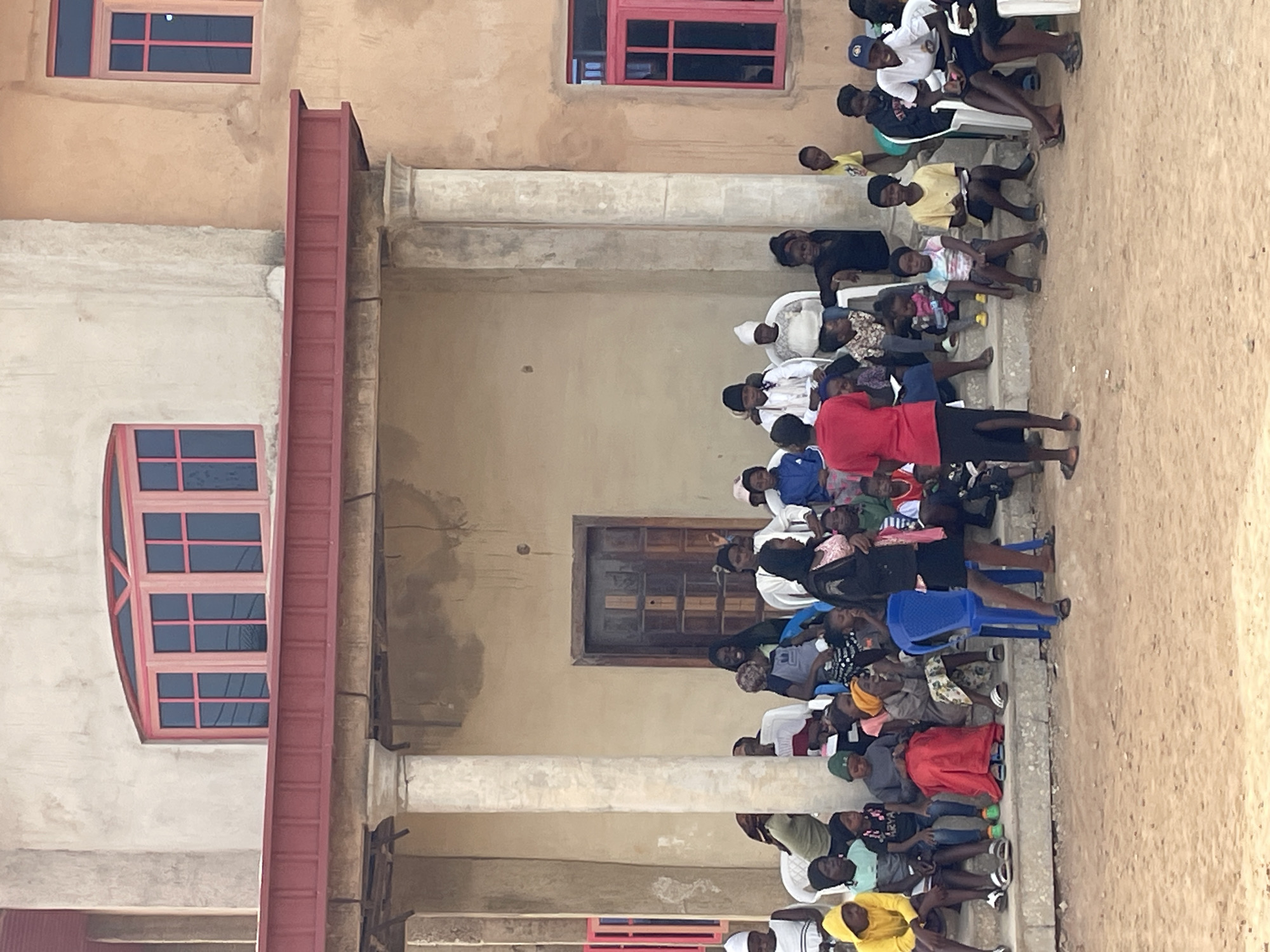 The image captures an outdoor gathering of a diverse group of people, primarily adults and children, in what appears to be a courtyard or open area next to a building in Jos, Nigeria. The scene is set during the daytime, under what seems to be clear and dry weather conditions, suggested by the even lighting and dusty ground.
A large number of individuals, predominantly of African descent, are present. Many are seated on white and blue plastic chairs, while others are on the ground. They are dressed in casual attire, including various shirts, t-shirts, dresses, and trousers, with some adults wearing head coverings. Numerous young children are interspersed within the crowd. One adult, distinctively wearing a red shirt and dark trousers, stands near the front of the gathering, facing the seated group, suggesting they are leading or addressing the assembly. The expressions on the faces of the attendees generally convey attentiveness and engagement.
The backdrop is formed by a multi-story building with light, possibly beige or tan, stucco-like walls. Concrete structural elements, including several stout pillars or columns, are visible, creating a partially shaded area where some of the people are gathered. The building features several windows with striking red frames and dark panes; one prominent window on the lower left is arched, while others are rectangular. A dark wooden door or window is also set into the building's facade. The ground beneath the gathering is a light brown, unpaved surface, likely dirt or sand. No visible text can be clearly read on any objects, clothing, or signs within the image.