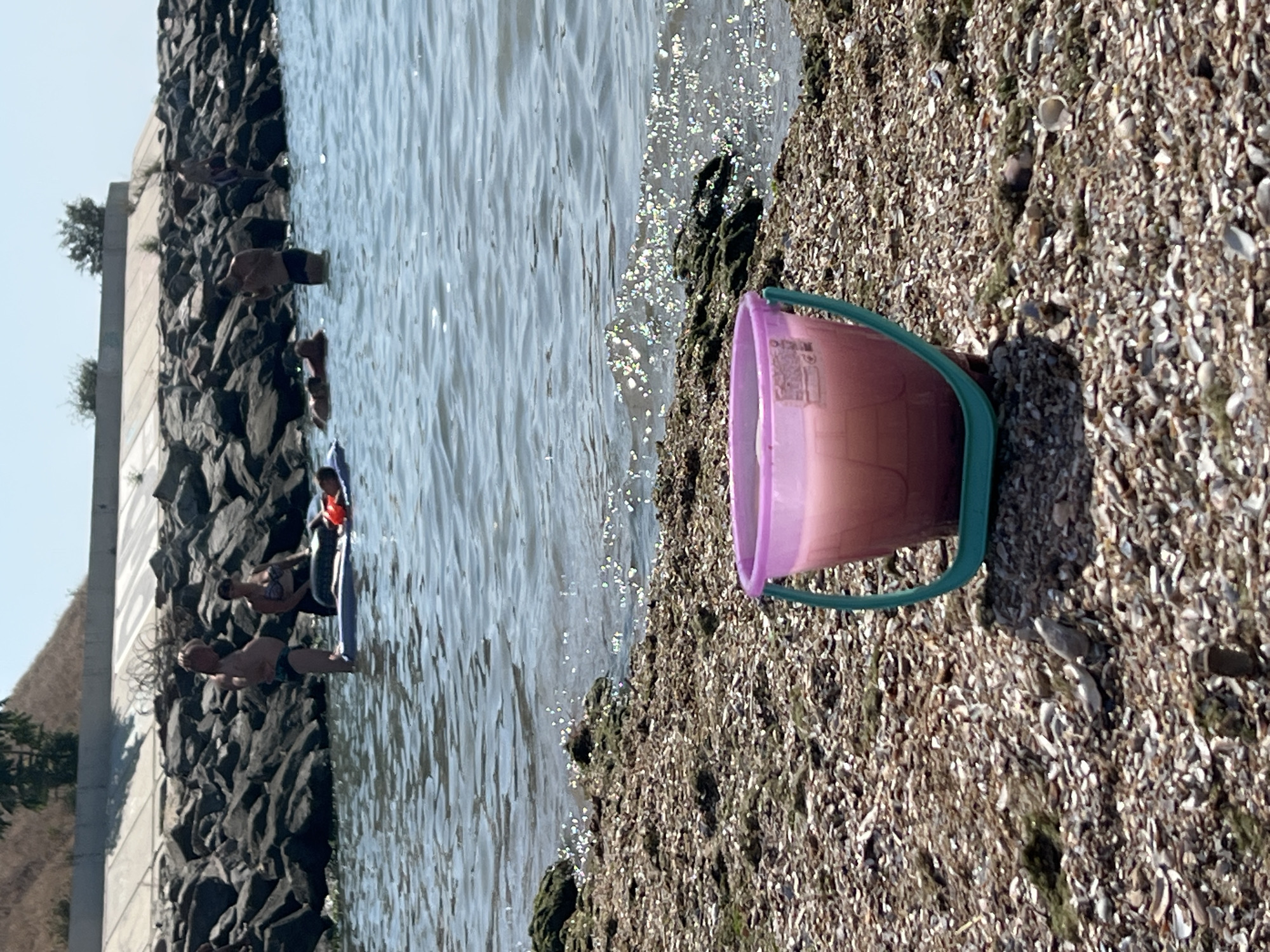 The image depicts a sunny day at the beach in Fontanka, Ukraine. In the foreground, a pink bucket with a teal handle sits on a pebbly shore covered with shells and seaweed. The bucket appears to contain a pinkish liquid. The water in the background is calm, with sunlight sparkling on its surface. Several people are visible in the water and on inflatable rafts. To the left, a man stands on a raft, and a woman sits behind him on the same raft. Further back, other individuals are seen in the water near a rocky breakwater that extends into the sea. The sky is clear and blue.