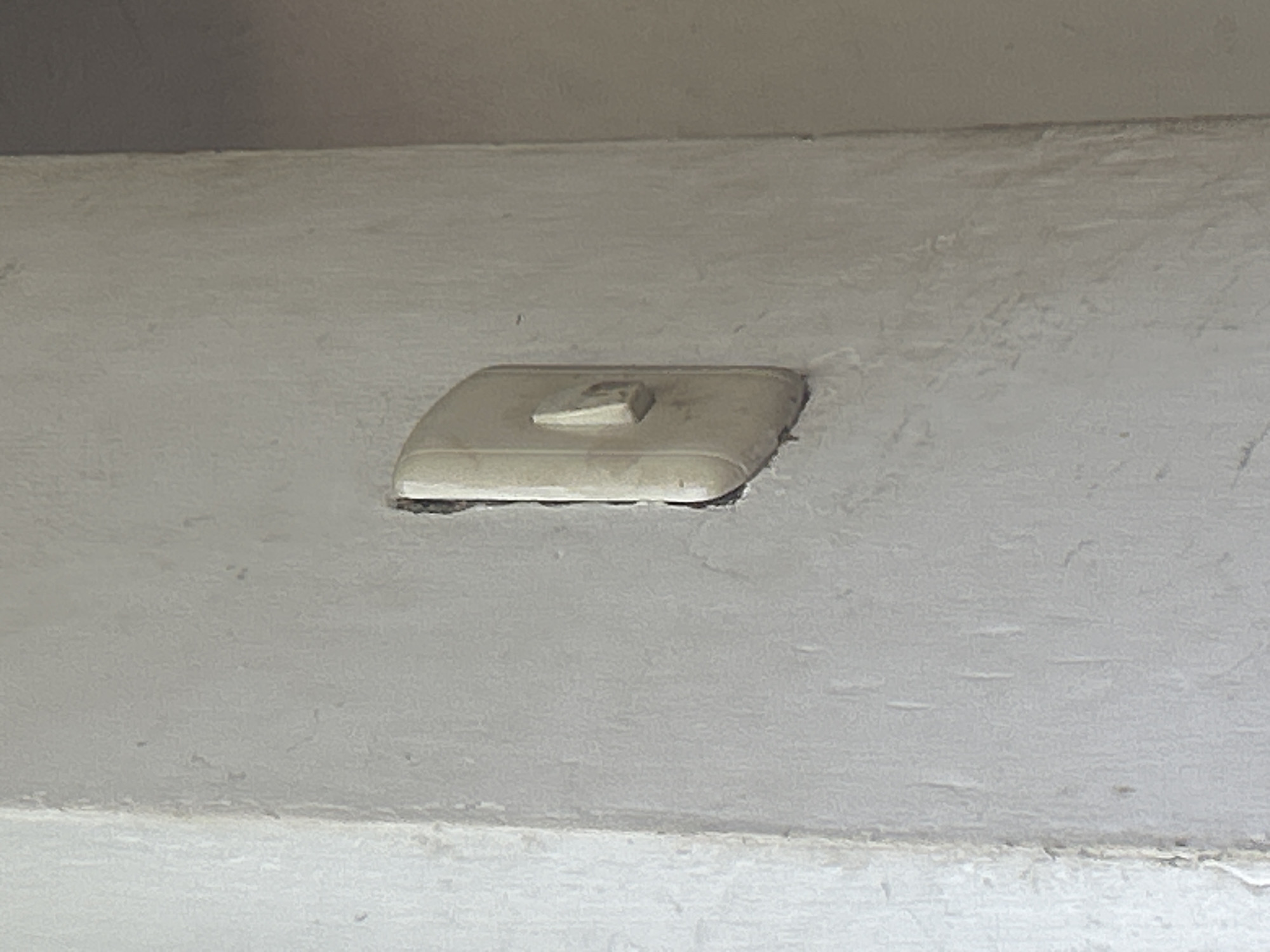 A close-up, slightly angled shot shows a weathered, off-white light switch embedded in a textured, off-white concrete wall. The light switch has a rectangular plate with a rounded top and bottom, and a small, triangular toggle protruding from its center. The wall surface is uneven, with visible brushstrokes and some minor discoloration, suggesting age or exposure to the elements. The image appears to be taken indoors, likely in an older building. The location is identified as Santa Ana, Venezuela.