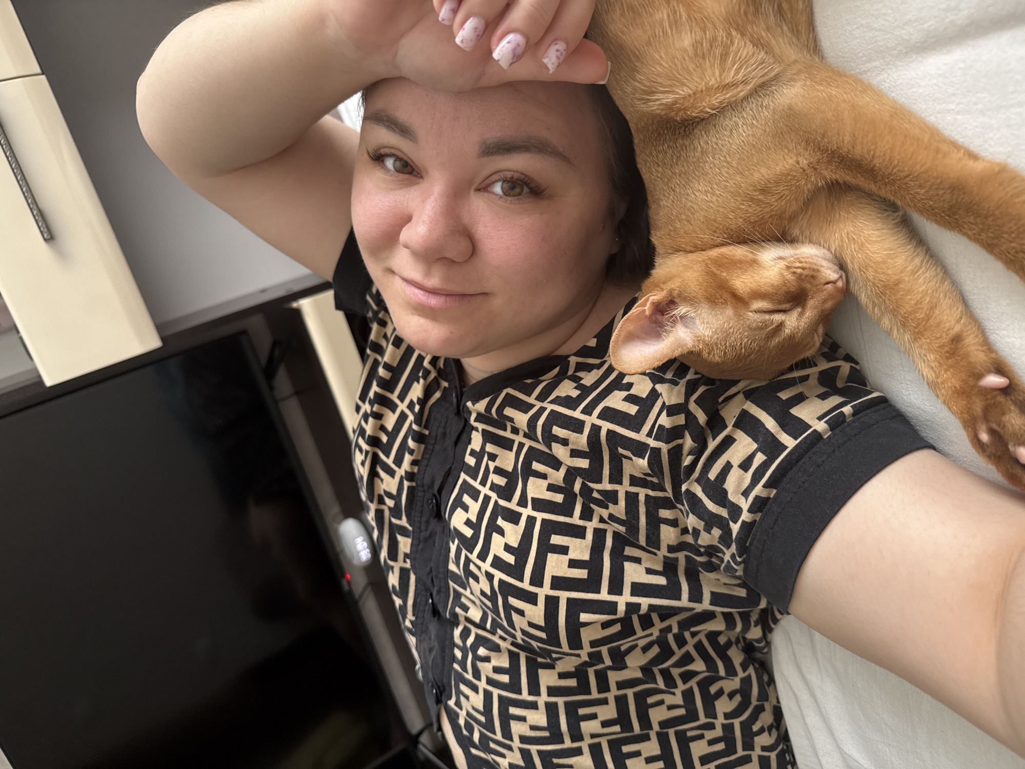 This is a close-up, overhead selfie of a woman with light skin and brown eyes lying down, with her right arm raised and bent over her forehead. She is smiling slightly and looking at the camera. A ginger cat is lying on her chest and shoulder, with its eyes closed and head resting on her shoulder. The cat appears to be sleeping.
The woman is wearing a black t-shirt with a repeating beige Fendi logo pattern. The cat is a light ginger color with subtle tabby markings. The background is slightly blurred, but appears to be a white surface, possibly a bed, and a dark, reflective surface to the left, likely a television screen. There are also some cabinets or furniture visible in the upper left corner.
The lighting suggests it could be daytime, as there is no indication of artificial lighting being the primary source. The scene is intimate and relaxed, conveying a sense of comfort and affection between the woman and her pet. No text is visible in the image. The location context is Stary Oskol, Russia.