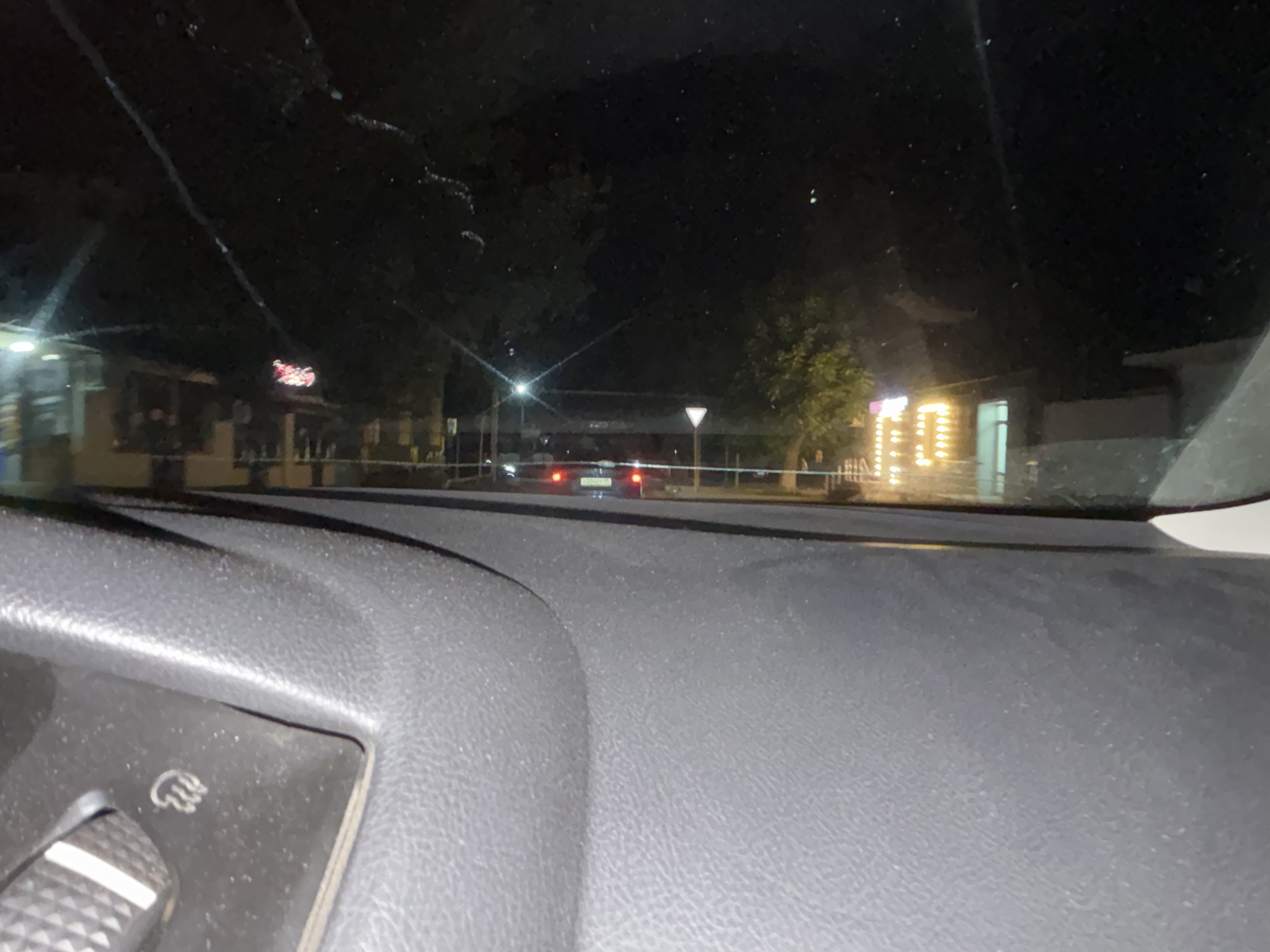 The image is taken from inside a vehicle at night, looking out through the windshield. The foreground shows the textured surface of a car's dashboard, with a partial view of a control knob. In the mid-ground, a street is visible with a few cars. One car in the center of the frame has its brake lights illuminated. There are trees and buildings lining the street, and some of the buildings have bright, illuminated windows or signs. A white and orange safety barrier is visible across the road in the mid-ground. The overall scene suggests a view of a street in the city of Grozny, Russia, at night.