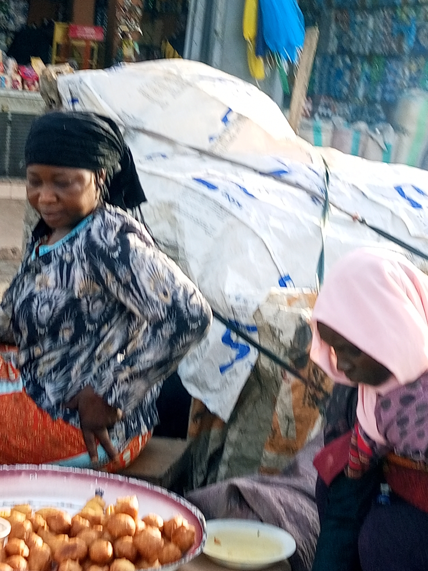 In Gombe, Nigeria, a street vendor with dark skin and black headscarf is captured from the chest up, showcasing a patterned black and grey blouse. Her gaze is directed downwards, her face partially obscured. Beside her, another person, also with dark skin, is visible from the chest up, wearing a pink headscarf and a dark purple garment adorned with a polka dot pattern. This individual's head is bowed, and their face is not visible. In the foreground, a large, oval-shaped platter holds a generous serving of fried dough balls, appearing golden brown and plentiful. The background is a blur of activity, featuring a large white tarp with blue markings, suggesting a bustling outdoor market environment, possibly near the Gombe main market.