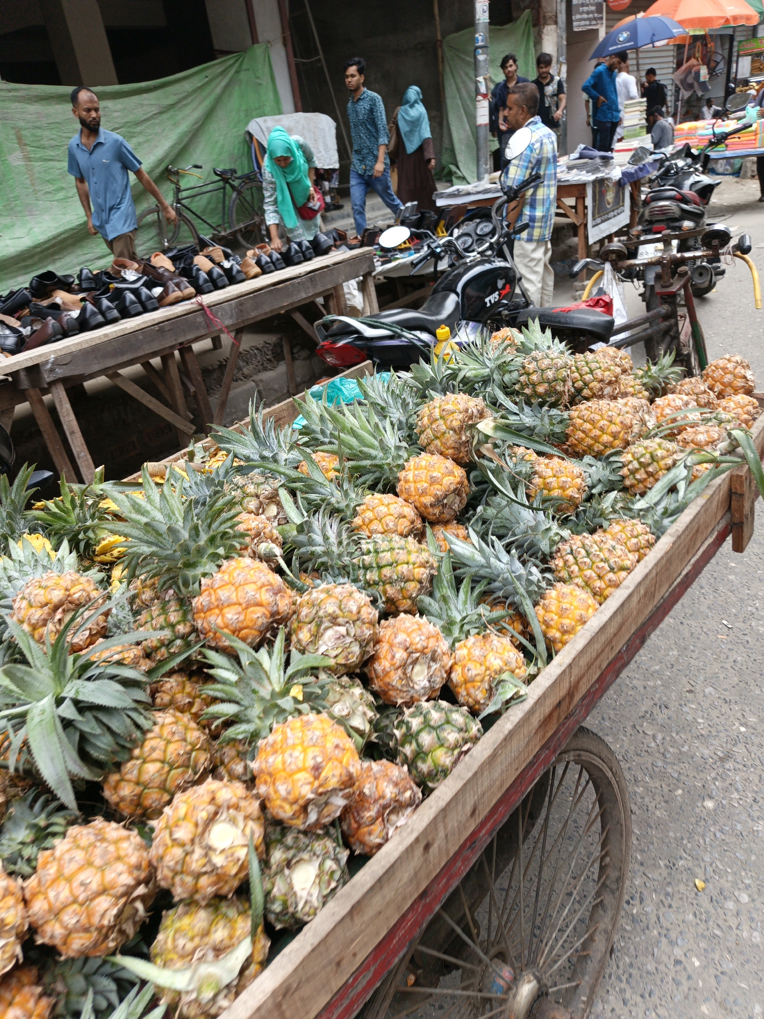 The image shows a street scene in Dhaka, Bangladesh, with a cart laden with pineapples in the foreground. The pineapples are small and orange-yellow, with spiky green tops. The cart is wooden with a single wheel. In the background, there are people walking, several motorcycles parked, and a man selling shoes and sandals on a wooden table. There is a green tarp hanging in the background. The scene is busy and crowded, typical of a marketplace or street vending area in Dhaka. The time of day appears to be daytime.