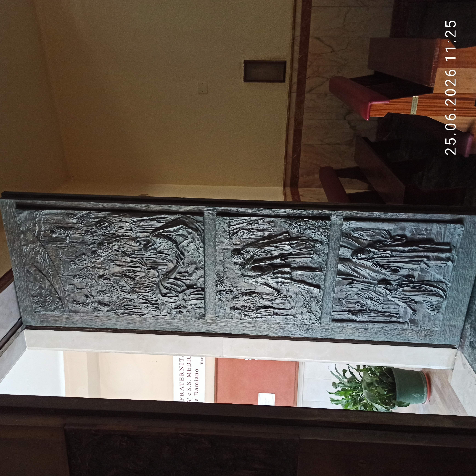 The image shows a close-up of a large, intricately carved bronze door, likely in a religious or historical building. The carvings depict scenes with multiple figures, appearing to be biblical or historical narratives, with detailed textures and flowing drapery. The door is divided into three panels, with each panel featuring a distinct scene.
To the right of the door, there are wooden benches with red cushioned seats, suggesting the setting is an interior space like a church, chapel, or monastery. The floor appears to be made of marble. A small plant in a dark green pot sits on the floor in the lower right portion of the image.
In the background, through the opening of the door, a sign with text is partially visible. The visible text reads "FRATERNITA' N' e S.S. MEDIC" and below that "e Damiano." The image also has a timestamp overlay in the upper right corner, reading "25.06.2026 11:25". The lighting suggests it is daytime, but the interior environment makes it difficult to determine the natural light conditions. The overall tone of the image is solemn and perhaps reverent, due to the nature of the door's carvings and the likely setting.