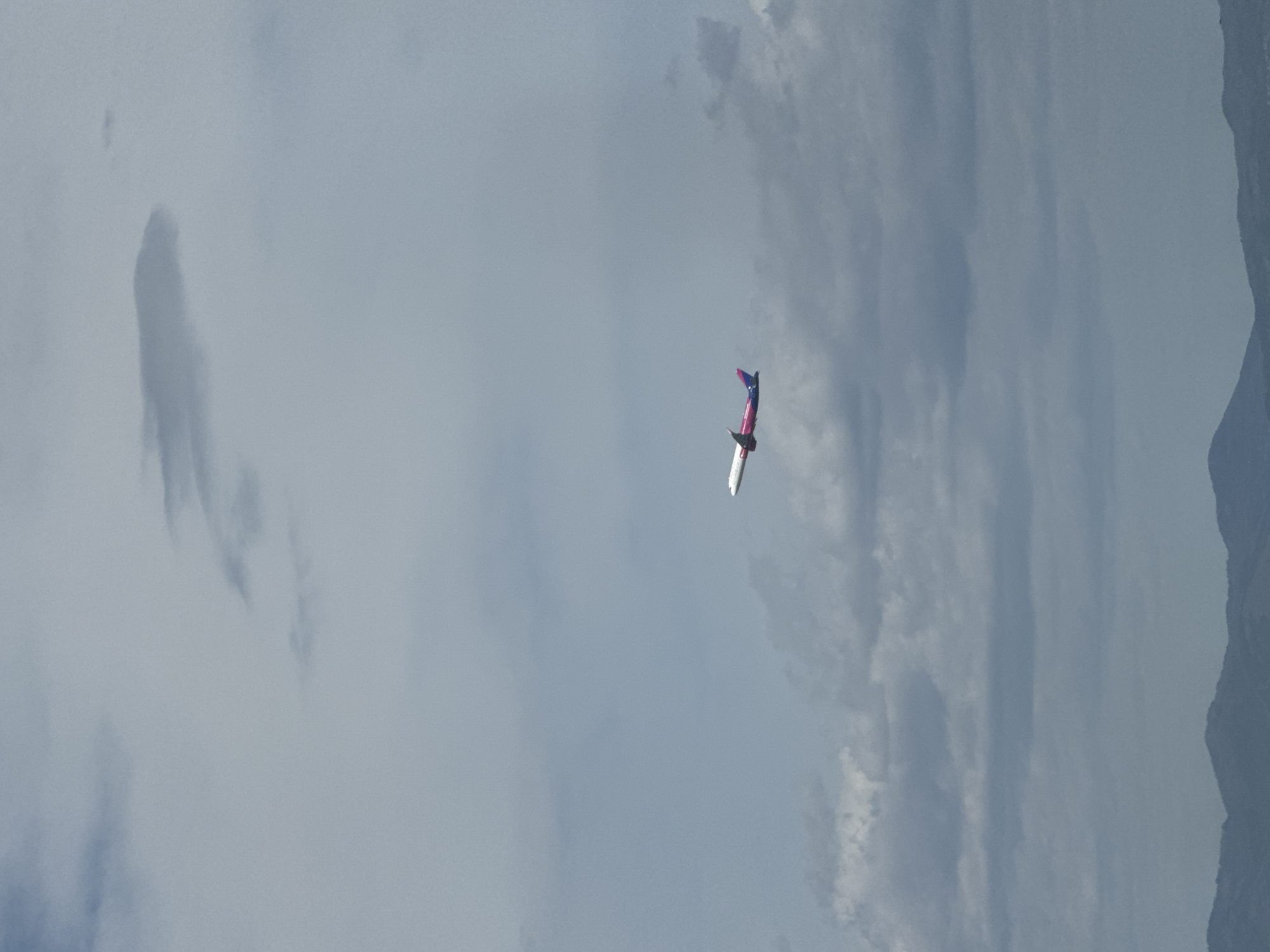 The image shows a plane in flight against a cloudy sky, possibly near a mountain range. The plane is red, white and blue. The location cannot be determined from the image to be in Sofia, Bulgaria.