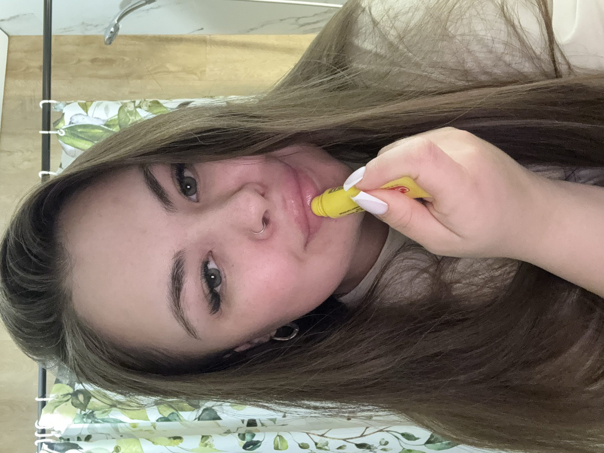 A woman is applying lip balm in what appears to be a bathroom. She has long brown hair, a nose ring, and is holding a yellow lip balm tube to her lips with her right hand, which has white manicured nails. The background includes a shower curtain with a floral pattern, a tiled wall, and part of a showerhead. The location in Murino, Russia is not discernible from the image.