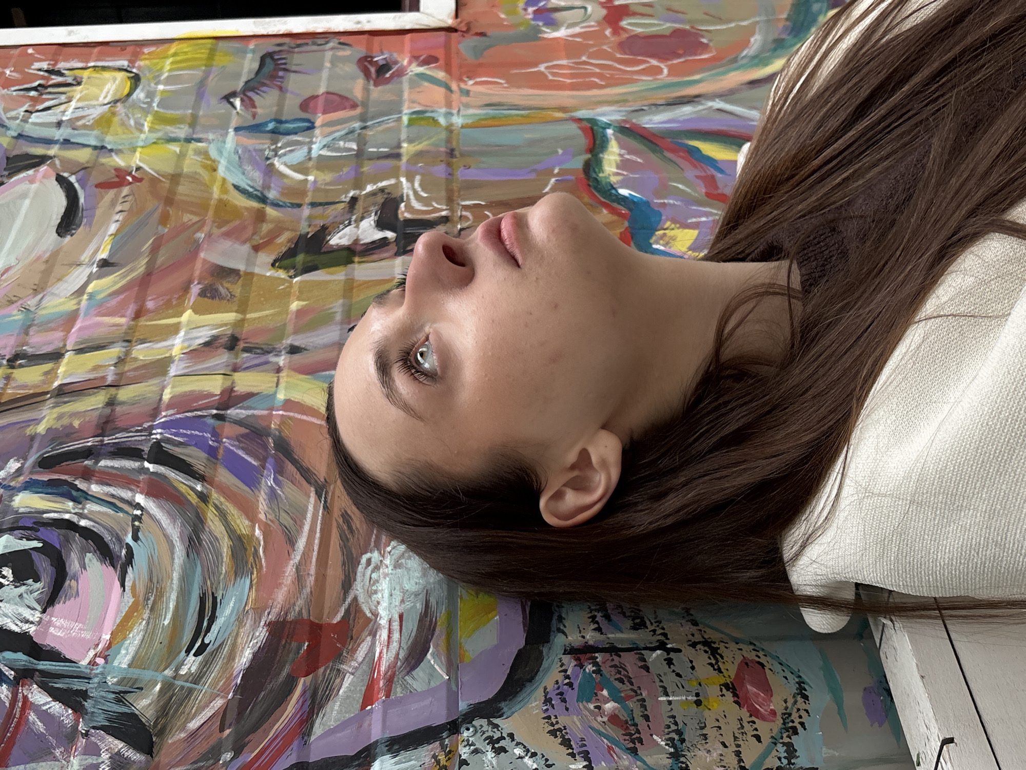 A medium, eye-level shot depicts a woman lying down, looking up and to her left. Her head rests against a painted surface, possibly a mural or large canvas, filled with swirls and strokes of various colors including orange, yellow, purple, and blue. The woman has light skin, brown hair, and distinctive green eyes. She appears to be wearing a white top. The location in the city of Kragujevac, Serbia, is not identifiable from the image.