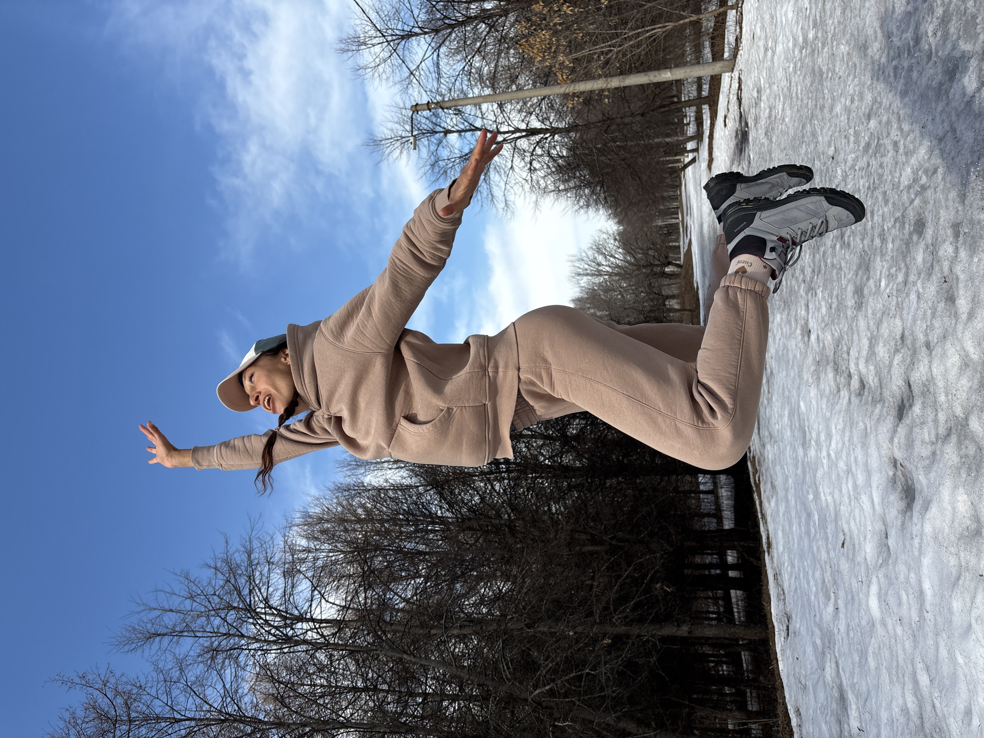 The image depicts a woman outdoors in a snowy environment, possibly in Ulyanovsk, Russia. She is wearing a beige tracksuit and grey hiking boots, with a baseball cap. She appears to be kneeling on the snow and has her arms outstretched. The background includes bare trees, some snow-covered ground, and a blue sky with clouds. A street lamp can be seen in the distance.