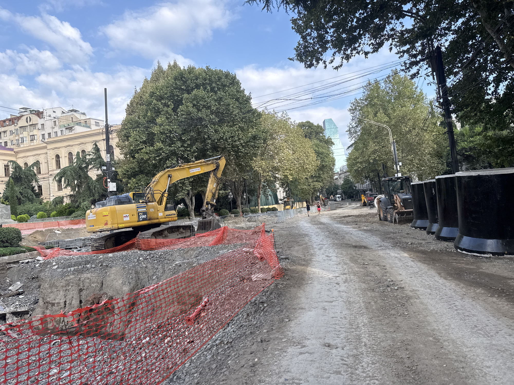 The image shows a construction site in Tbilisi, Georgia, characterized by a dirt road, an excavator, and various construction materials. In the background, a distinctive modern skyscraper with a greenish-blue facade stands tall, identifiable as the "Georgia's Parliament Building" or the "House of Justice" in Tbilisi, which is a prominent landmark. The foreground is dominated by an excavator, likely a Komatsu PC290, positioned next to a deep excavation pit cordoned off by orange safety netting. Scattered debris and piles of gravel are visible around the pit. The road itself is unpaved, appearing to be undergoing construction or repair, with tire tracks indicating recent activity by heavy machinery. Several large, black, cylindrical objects are lined up along the right side of the road, potentially part of the ongoing construction. Two individuals are walking away from the camera on the dirt road in the mid-ground, their presence suggesting ongoing activity despite the construction. The sky is partly cloudy, with a few wispy clouds against a blue backdrop. Lush green trees line the sides of the road, with some foliage from the trees also visible in the foreground. A large, classical-style building, likely a government or public institution, is situated to the left of the excavator, partially obscured by trees. Overhead power lines are visible crossing the scene, typical of urban infrastructure.