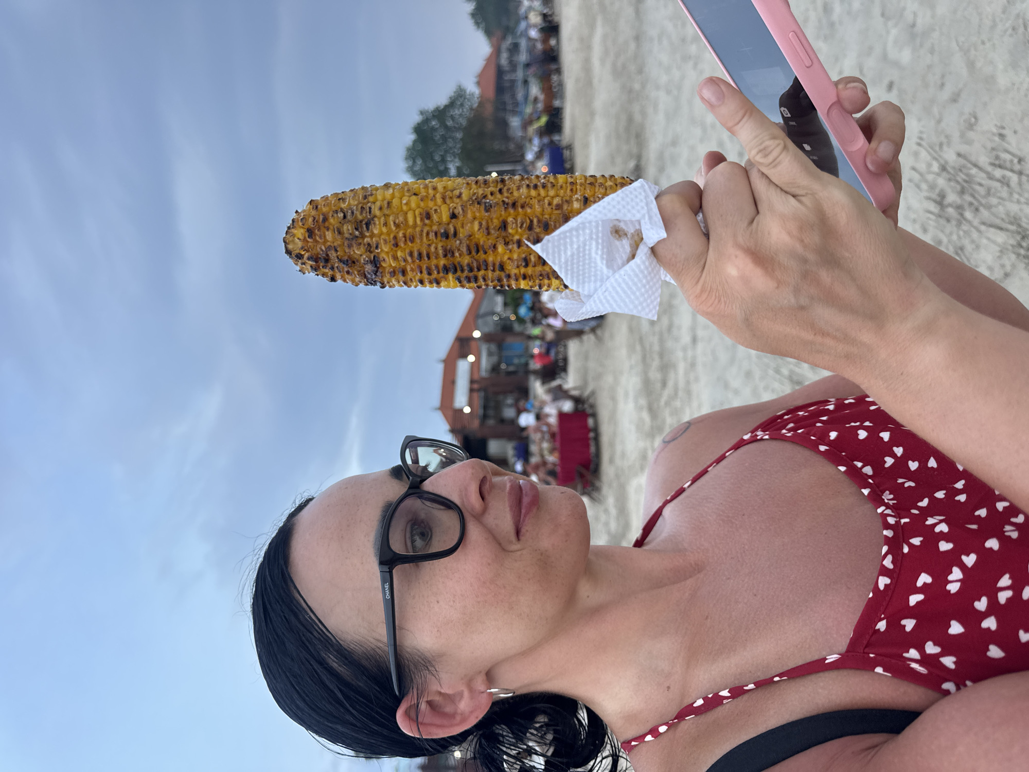 A woman with dark hair pulled back, wearing black-framed "CHANEL" glasses, silver hoop earrings, and a red halter-neck top patterned with white hearts, stands on a sandy beach in Jimbaran, Indonesia. A faint, circular tattoo is visible on her right shoulder. In her left hand, she holds a large, char-grilled ear of corn partially wrapped in a crumpled white paper napkin. Her right hand operates a smartphone in a pink case; a dark reflection is visible on its screen. Her gaze is directed towards the right. In the mid-ground, behind the woman, are multiple open-air restaurant structures with traditional red-tiled roofs. Numerous blurred figures are seated at tables in front of these establishments, some with red tablecloths. Green vegetation is visible beyond the structures. The sky overhead is overcast and light blue-grey, consistent with late afternoon or early evening lighting.
