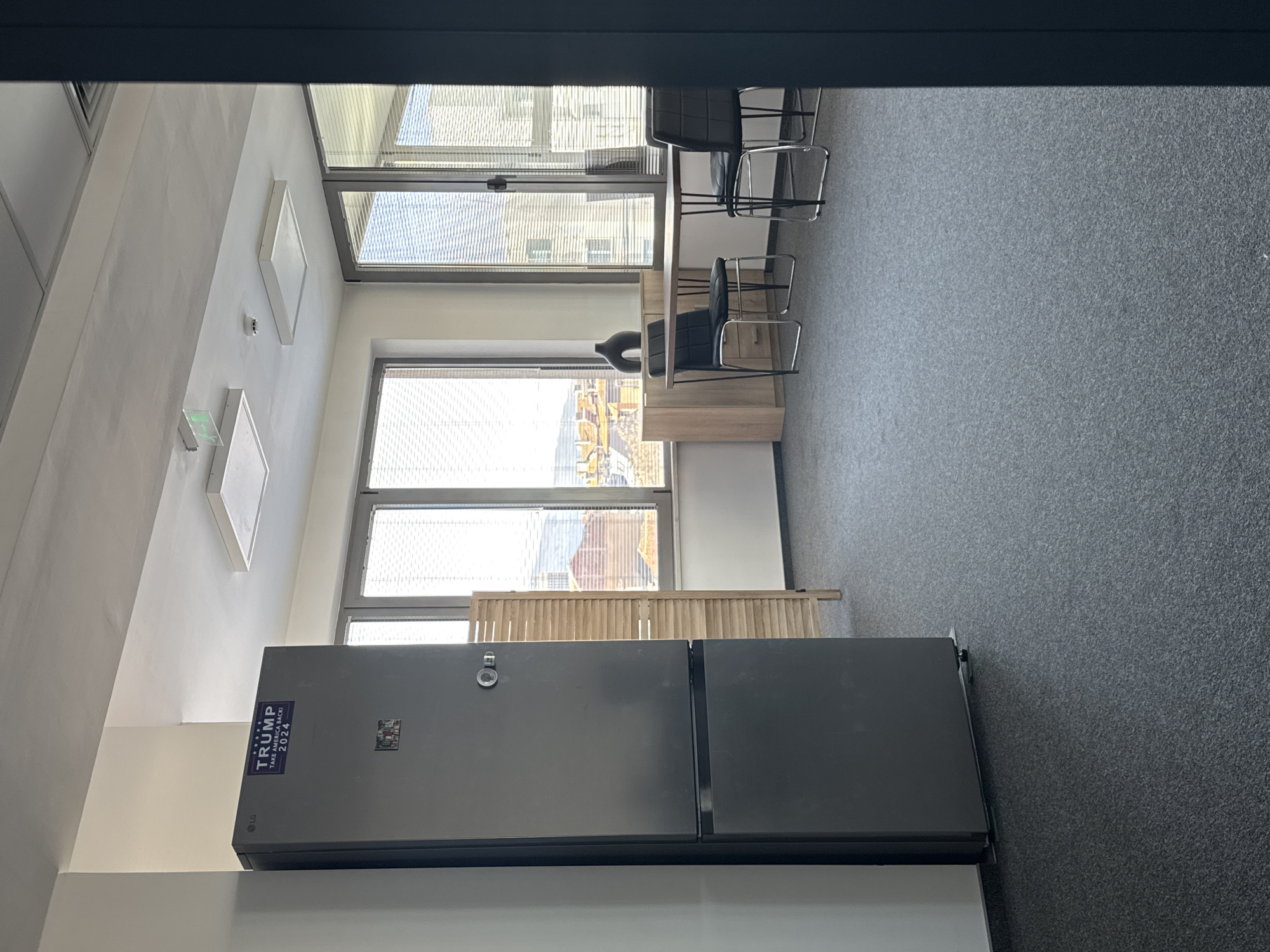 The image depicts an interior view of a modern office space, potentially in Sofia, Bulgaria, as suggested by the urban landscape visible through the windows. The room is characterized by abundant natural light streaming in through multiple large windows, some of which are equipped with blinds. The flooring is a uniform gray carpet. The room's furnishings include a light-colored desk with black office chairs, and a large, modern refrigerator with a "TRUMP - TAKE AMERICA BACK! 2024" sticker on it. There are also some decorative elements like the wooden screen and wall-mounted lighting fixtures. There are no people visible in the frame, and the focus is on the architectural and interior elements of the office space.