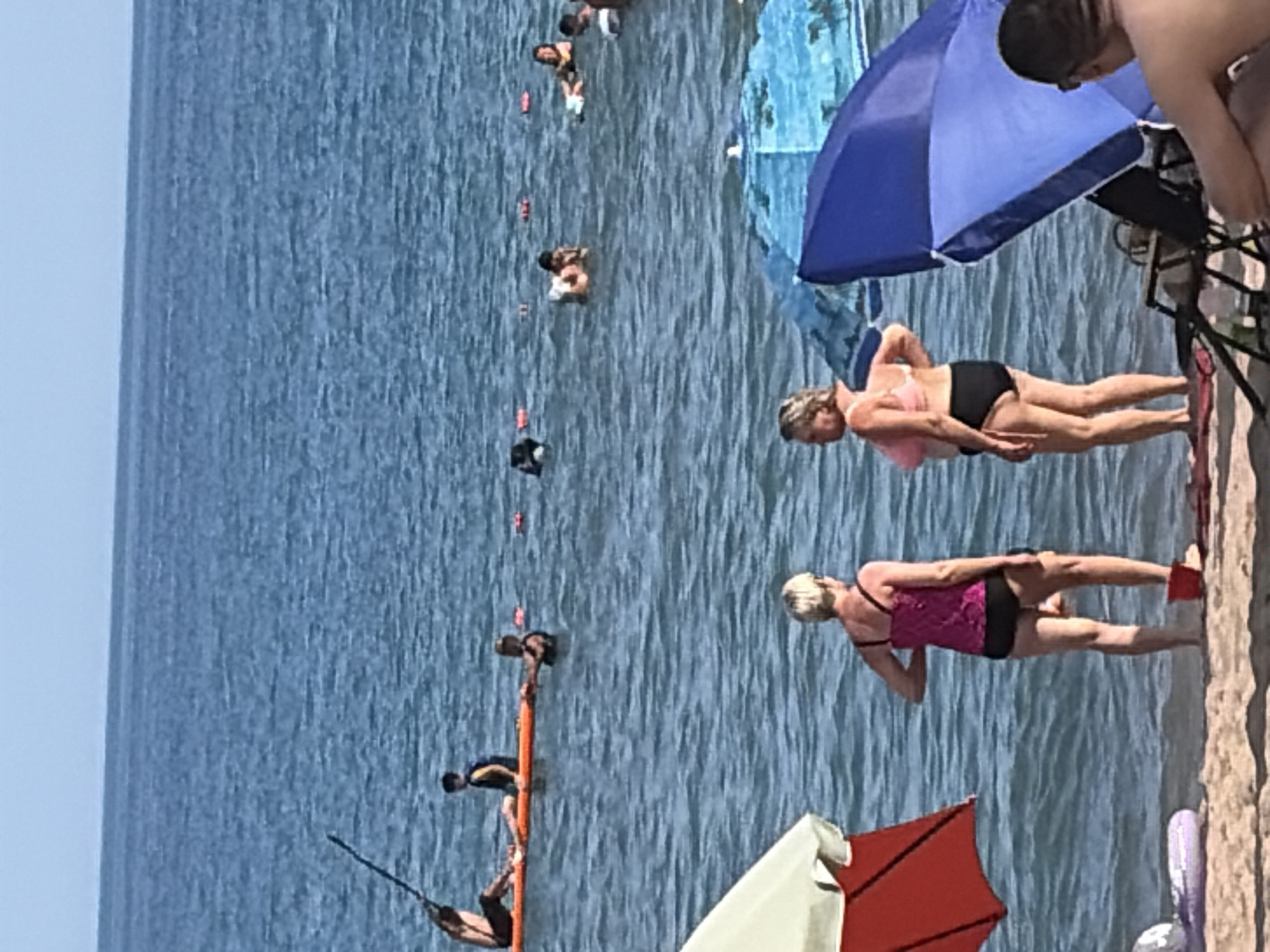 In a full, distant shot of a beach scene, two women stand in the shallow water near the shoreline. The woman on the left wears a pink tank top and black bottoms, her back to the camera. The woman on the right, also facing away, wears a pink bikini top and black bottoms. In the background, several people are visible in the ocean. Some are swimming, while others are on or near a paddleboard. A paddleboarder with a black pole stands on a paddleboard. Further out, a group of people are in the water, some on inflatable devices. The water is blue and calm, with a clear horizon line separating it from a pale blue sky. Beach umbrellas and chairs are partially visible on the sand in the foreground.
