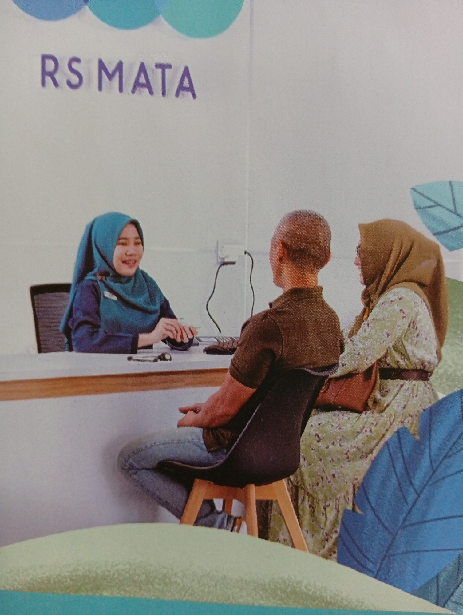In the city of Tlogomulyo, Indonesia, an image captured within the RS Mata, a medical facility, shows a reception area. A female employee, wearing a teal hijab and uniform, is seated behind a counter, smiling as she operates a computer mouse and keyboard. Two customers, a man and a woman both wearing hijabs, are seated in front of the counter, facing the employee. The man is wearing a brown polo shirt and jeans, while the woman is in a green floral dress. A brown handbag rests on the woman's lap. The environment appears clean and modern, with a light-colored counter and white walls adorned with abstract blue and green leaf motifs and the "RS MATA" logo.