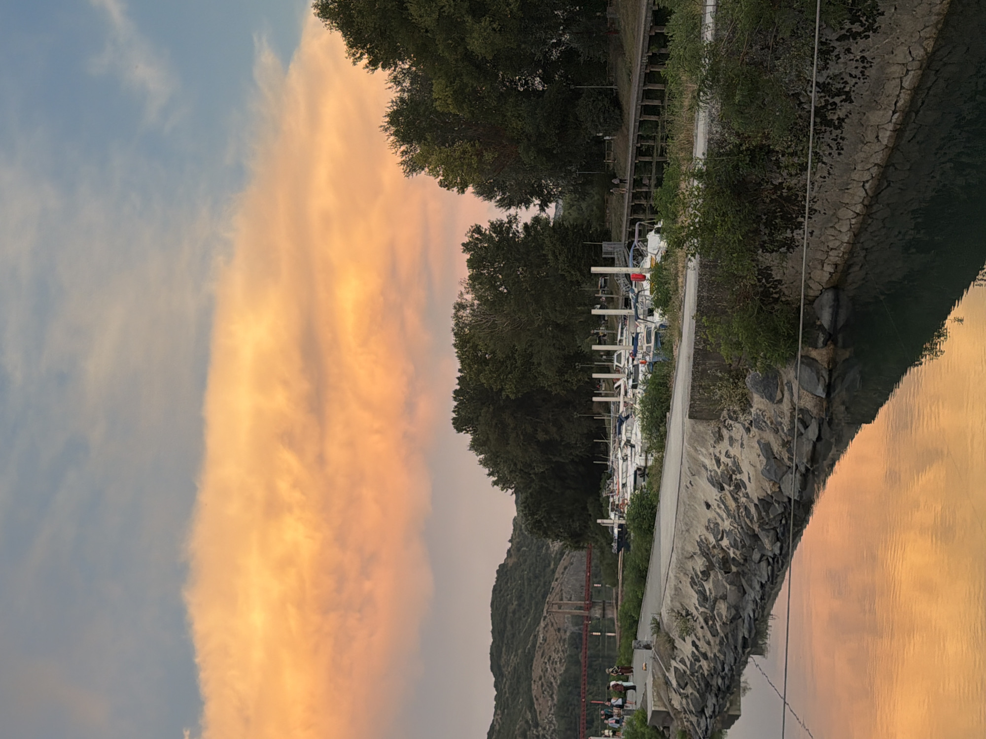 The image depicts a serene waterside scene in Viviers, France, under a vibrant sunset sky. The upper portion of the frame is dominated by a sky filled with fluffy clouds painted in hues of orange and pink, transitioning to a soft blue. Below the sky, a line of lush green trees forms a dense backdrop.
In the mid-ground, a marina is visible, lined with numerous white boats docked neatly. A wooden walkway or pier runs along the waterfront, with a few individuals strolling or standing, appearing to observe the scenery.
The foreground features a stone-lined embankment leading to the water. The water itself is calm, reflecting the colorful sky and the surrounding elements, creating a mirror-like effect. In the distance, across the water, a red suspension bridge is visible, connecting two hills. The hills are covered in vegetation and appear rugged.
The overall impression is one of tranquility and natural beauty, capturing the essence of a peaceful evening by the water in a French locale.