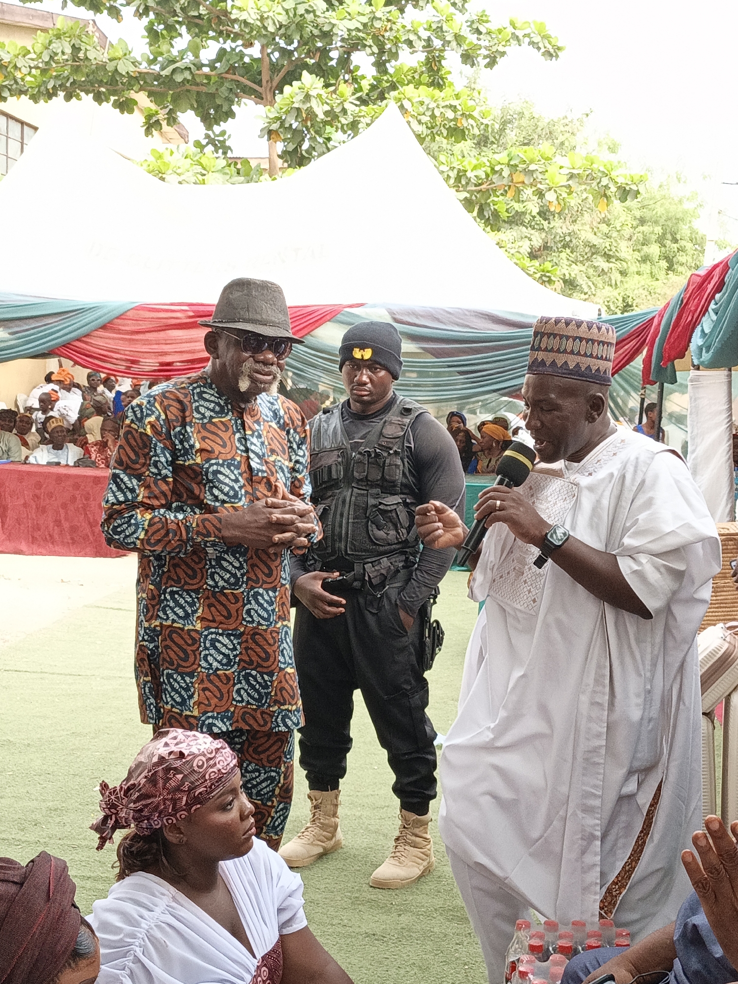 This daytime image captures an outdoor public gathering or ceremony in Gawuna, Nigeria, with a focus on three prominent men and a woman in the foreground. The scene is set under white tents adorned with drapes in red, green, and blue fabric, over a green artificial turf. Lush green trees are visible in the background, suggesting a clear day.
On the left, an older man stands, wearing a dark fedora, sunglasses, and a colorful patterned traditional long-sleeved shirt (possibly an Adire or Ankara print) with shades of brown, blue, and black. He has a distinctive white beard and mustache and his hands are clasped in front of him. Next to him, a younger man, likely security personnel, stands in a black beanie with a yellow logo, dark tactical vest, dark long-sleeved shirt, black pants, and light-colored boots. He maintains a serious expression.
To the right, a man in a flowing white traditional garment (agbada) with subtle gold embroidery on the chest and a multi-patterned, embroidered cap (fila) holds a microphone and appears to be speaking or singing. He wears a watch on his left wrist.
In the bottom left foreground, a woman in a white top and a patterned reddish-brown headscarf sits on the ground, looking intently to her left. Other attendees are visible in the background, seated under the tents, many also in traditional attire. A crate of bottled beverages is seen on the ground near the speaker. No discernible text is clearly visible on any banners, clothing, or objects.
