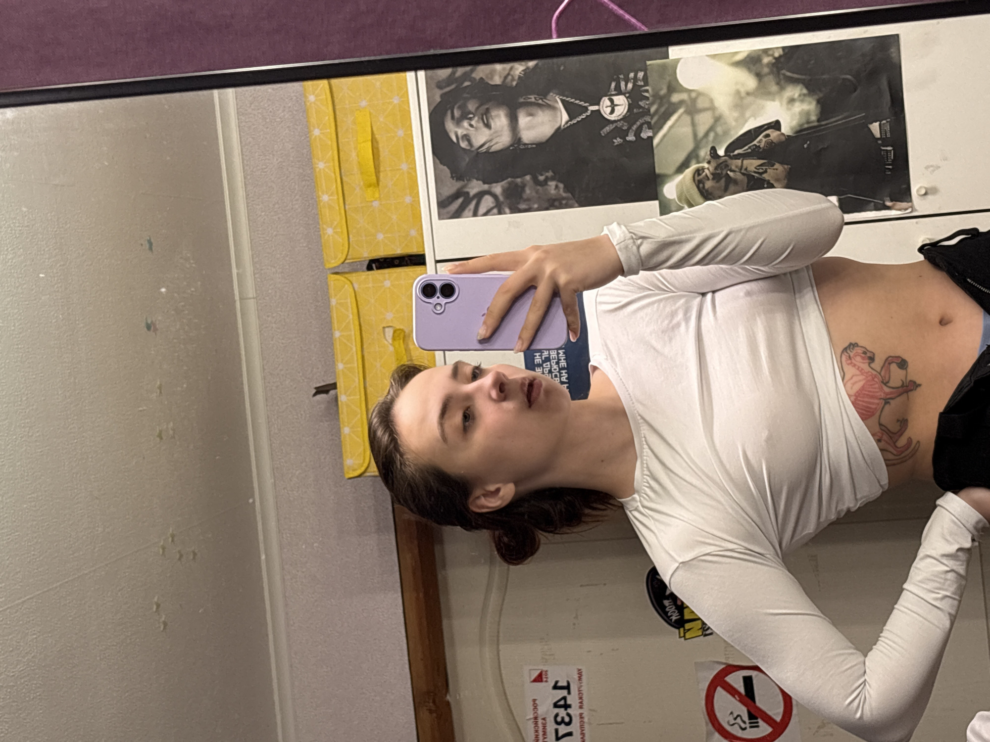A woman is taking a mirror selfie in an indoor space. She is wearing a white long-sleeved crop top and black bottoms, revealing a tattoo on her stomach. She is holding a purple smartphone. The background includes yellow storage cubes, two framed posters (one with musicians), and wall stickers including a "no smoking" symbol and writing in Russian, potentially indicating a location in Прикамский, Russia. The room appears to be a dressing room or a personal space.