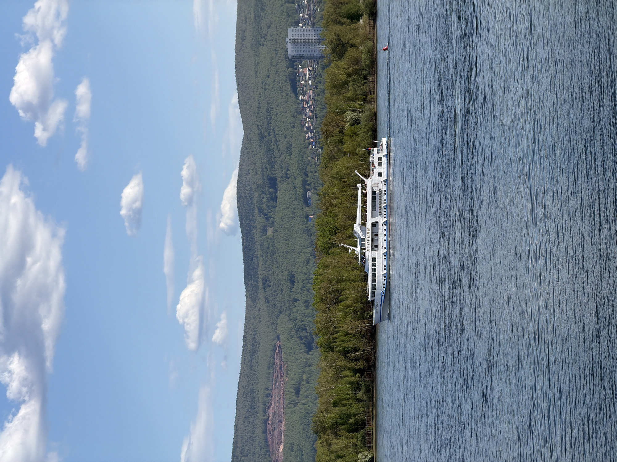 The image shows a sideways view of the Yenisei River in Krasnoyarsk, Russia, with a white passenger ship sailing on the water. The sky is blue with scattered clouds. A steep, forested mountain range, likely part of the Stolby Nature Reserve, runs along the edge of the riverbank. Buildings are visible along the mountain's edge. A red buoy floats near the ship. The Yenisei River's waters are visible.
