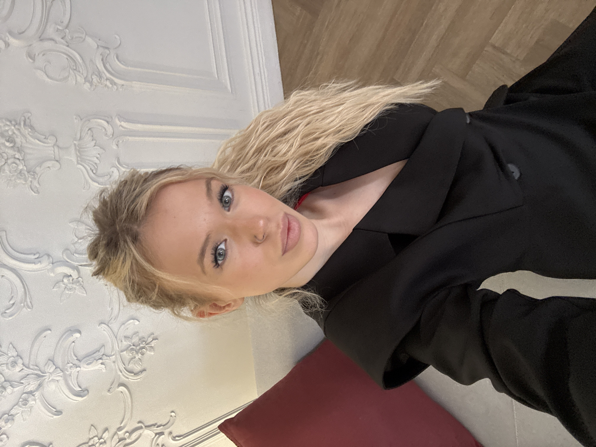 A medium shot selfie captures a blonde woman from the chest up. She is indoors, seated with a burgundy cushion to her left and a light-colored sofa beneath her. Her hair is styled in a high ponytail, and she wears a black blazer with a red garment visible underneath. The background features ornate white decorative molding on the ceiling and a light wooden floor. The overall impression is one of an interior setting, possibly a room with classical architectural elements. The image does not contain any discernible information to place it specifically within Saint Petersburg, Russia.