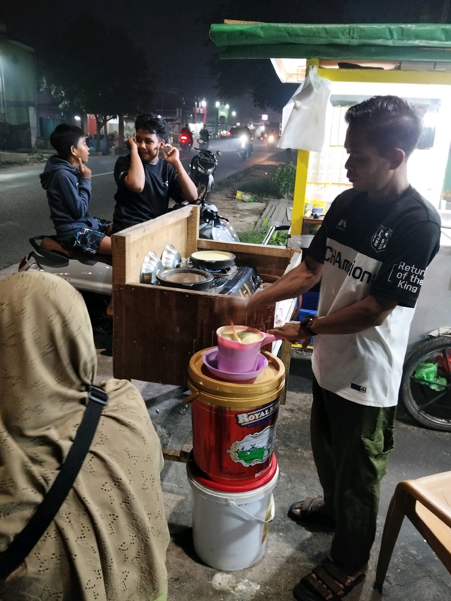 The image depicts a street food stall at night in Arang Limbung, Indonesia. A vendor, wearing a black and white t-shirt and green pants, is actively preparing food. He is holding a pink cup filled with a green liquid, possibly a beverage or dessert, and appears to be stirring it. In front of him is a wooden food stall structure with various cooking utensils and containers, including pots and bowls. Stacked on the ground next to the stall are several large plastic buckets, one red and one white.
To the left of the vendor, two young boys are seated on a motorcycle. One boy, wearing a blue hoodie and patterned shorts, is looking towards the vendor. The other boy, in a black t-shirt, is looking down and touching his ears. A woman wearing a light brown headscarf and a black strap is partially visible in the foreground, facing away from the camera and towards the stall.
The background shows a street scene at night with illuminated buildings, streetlights, and passing vehicles. The overall atmosphere suggests a busy urban environment with ongoing food commerce.