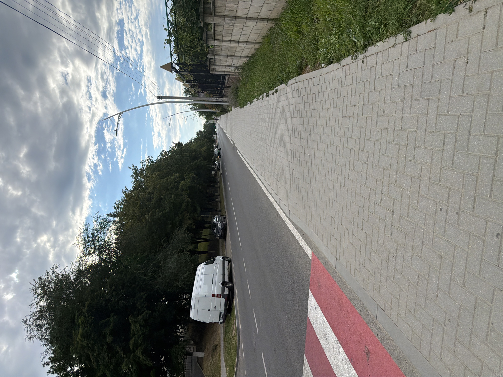 A street scene in Strășeni, Moldova, depicts a wide asphalt road with a pedestrian sidewalk. On the left side of the road, a white van is parked, and a crosswalk with red and white stripes is visible. Trees line the left side of the road, and on the right, a concrete wall separates the sidewalk from a grassy area and vegetation. Several streetlights are positioned along the sidewalk, and power lines stretch across the sky. The sky is partly cloudy, with patches of blue sky visible between the clouds. The image is captured from a low angle, looking down the length of the street.
