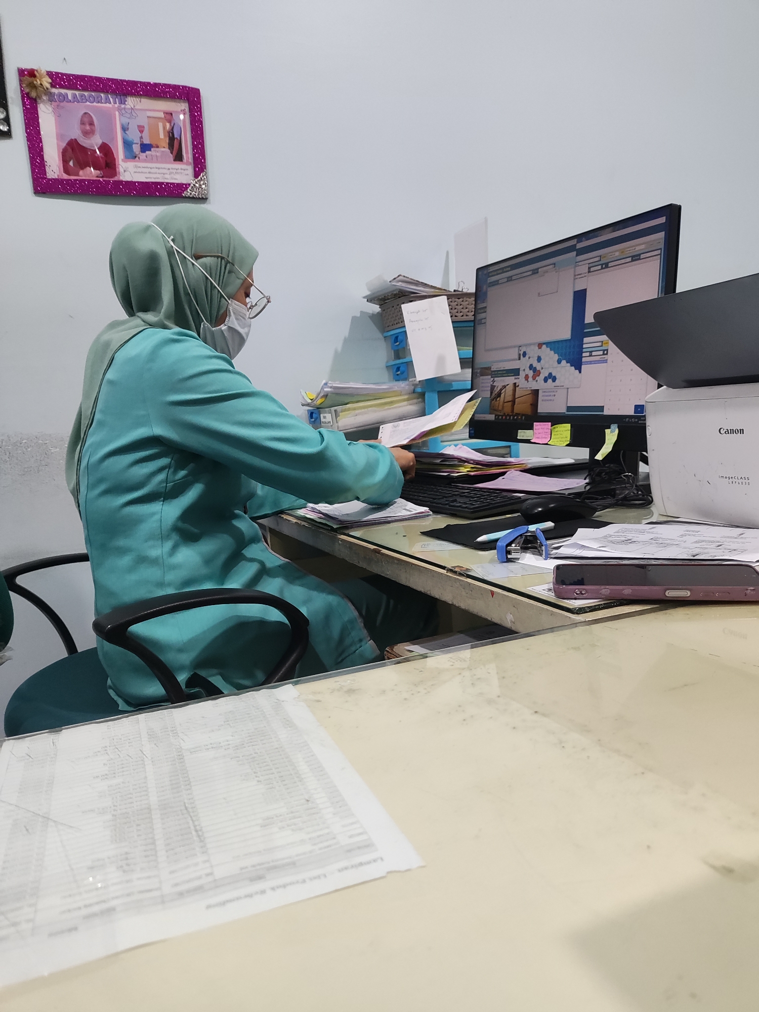 An indoor scene depicts a person wearing a teal scrub suit, a light green hijab, glasses, and a white surgical mask. The person is seated at a desk in what appears to be an office environment, with their right hand engaged with papers on the desk. A computer monitor displaying various windows and data is positioned behind the person. A Canon ImageCLASS printer is visible to the right of the monitor. The desk is cluttered with papers, folders, a stapler, a blue pen, a mouse, and a smartphone. In the background, a framed picture hangs on a light blue wall, showing two images: one of a woman in a red shirt and another of two people in medical attire. The overall environment suggests a workspace, possibly in a medical or administrative setting within Surabaya, Indonesia.