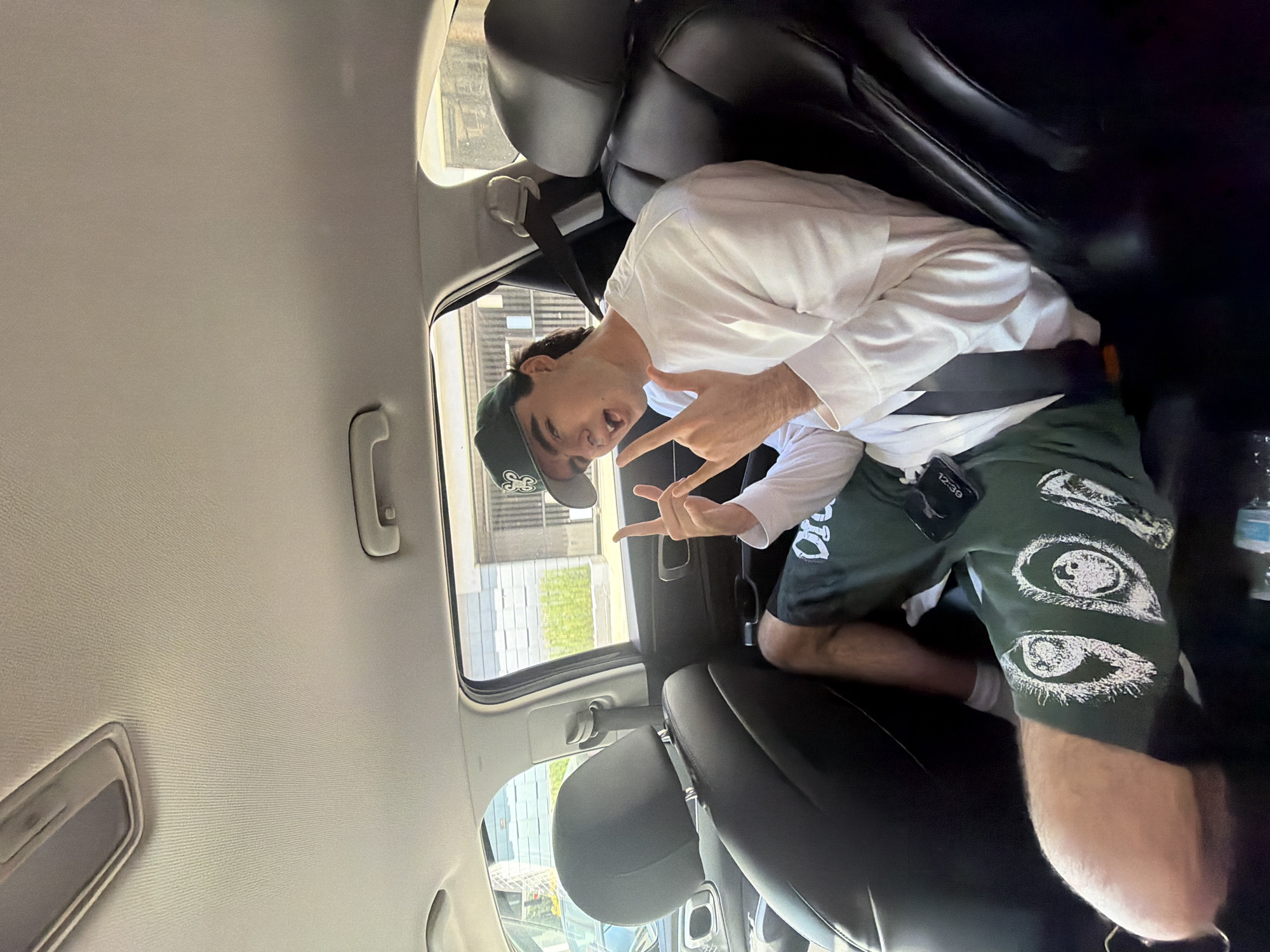A male wearing a white long-sleeve shirt, green shorts with eye graphics, and a green baseball cap sits in the back seat of a car, making a peace sign with both hands. The time on the digital device attached to his shorts reads 12:39. The view through the car windows shows a building with green vegetation and what appears to be an adjacent street or sidewalk. The location is in the city of West Hollywood, United States of America.