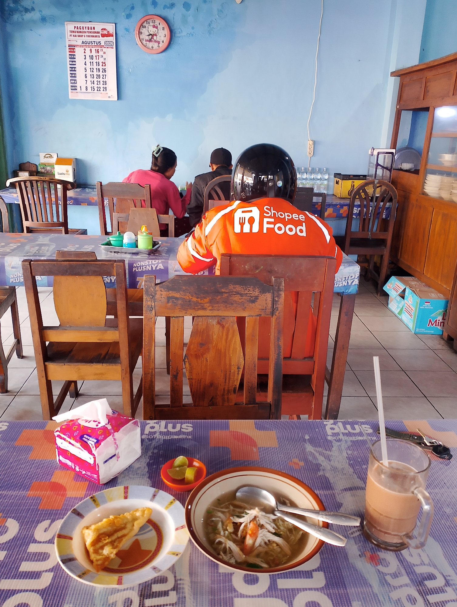 The image depicts an indoor dining area in what appears to be a casual eatery in Sinduadi, Indonesia. The overall ambiance suggests a local restaurant or food stall. In the foreground, a table is set with a meal, including a bowl of soup, a fried pastry on a plate, lime wedges in a small dish, and a glass of iced coffee with a straw. A box of tissues is also present on the table. The table is covered with a patterned plastic tablecloth.
In the midground, several wooden chairs are arranged around tables. A person wearing an orange Shopee Food jacket and a black helmet is seated, facing away from the camera. Two other individuals, a woman in a pink shirt and a man in a dark jacket, are also seated at a table, facing away from the camera. The wall behind them is painted a light blue and features a calendar for August 2026 and a pink cat-themed clock.
In the background, a wooden cabinet with glass doors is visible, filled with dishes. On the floor, several cases of bottled water are stacked. The environment appears somewhat utilitarian, with a tiled floor and simple furnishings. The lighting is natural and slightly diffused, suggesting it might be daytime. The presence of the Shopee Food jacket indicates a food delivery service operating in the area.