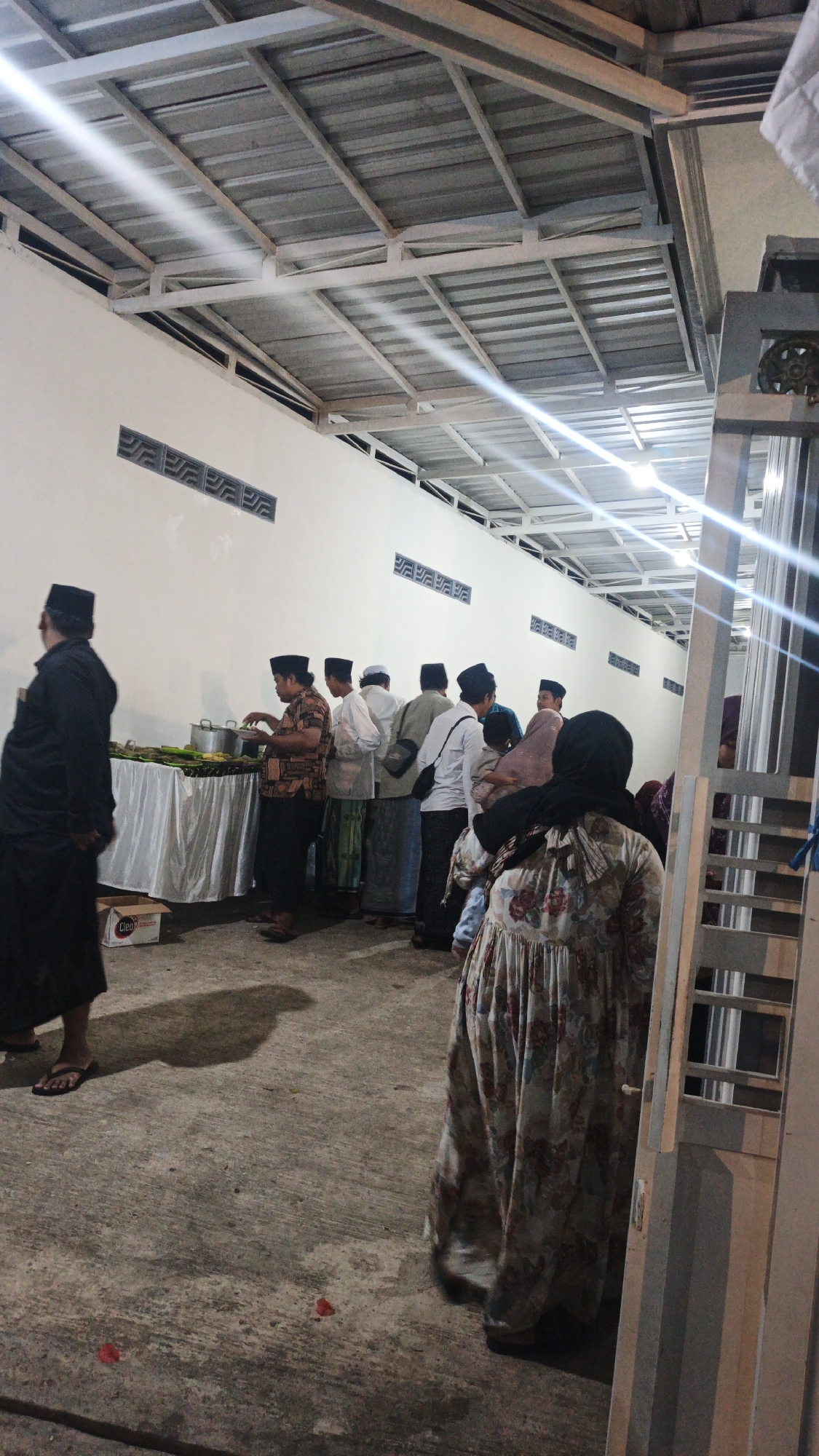 In Pandanarum, Indonesia, a group of people are gathered outdoors under a corrugated metal roof supported by a white metal frame. The scene appears to be a communal event, possibly a meal, with a long table covered by a white cloth displaying what appears to be food wrapped in banana leaves and some bowls. Several men, most wearing peci hats and sarongs, are standing in line, some reaching for food from the table. One man in the foreground, on the left, is dressed in black and facing away from the camera. Further back and to the right, a woman wearing a floral dress and hijab is also visible. The environment is a concrete ground with some scattered red petals. The wall in the background is white with decorative vent-like patterns. The lighting suggests it is either evening or an overcast day, with light fixtures visible on the ceiling.