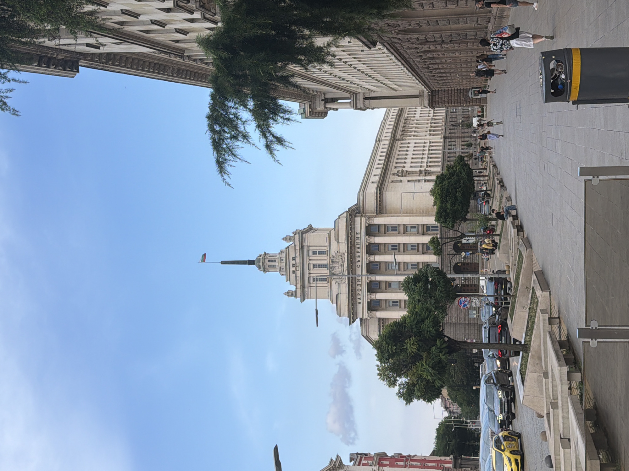 The image depicts a wide shot of a grand, light-colored building with classical architectural elements, including columns and ornate detailing. A flagpole with a small flag is visible at the top of a spire on the building. The building's facade displays the text "НАРОДНО СЪБРАНИЕ" which translates to "National Assembly" in Bulgarian, identifying the location as the Bulgarian Parliament in Sofia.
To the right of the building, a paved walkway lined with planters and trees runs alongside a street with a line of parked cars. Several people are walking on the sidewalk, some appearing to be tourists, while others seem to be local residents. One person is carrying a backpack, and another is wearing a bright floral shirt. A large, grey trash bin with a yellow stripe is prominently placed on the sidewalk.
The scene is bathed in daylight, suggesting it is daytime. The sky is mostly clear with a few scattered clouds. The overall atmosphere appears to be calm and urban. The perspective is slightly tilted, giving a dynamic feel to the composition. The architecture of the building suggests a historical and important site.