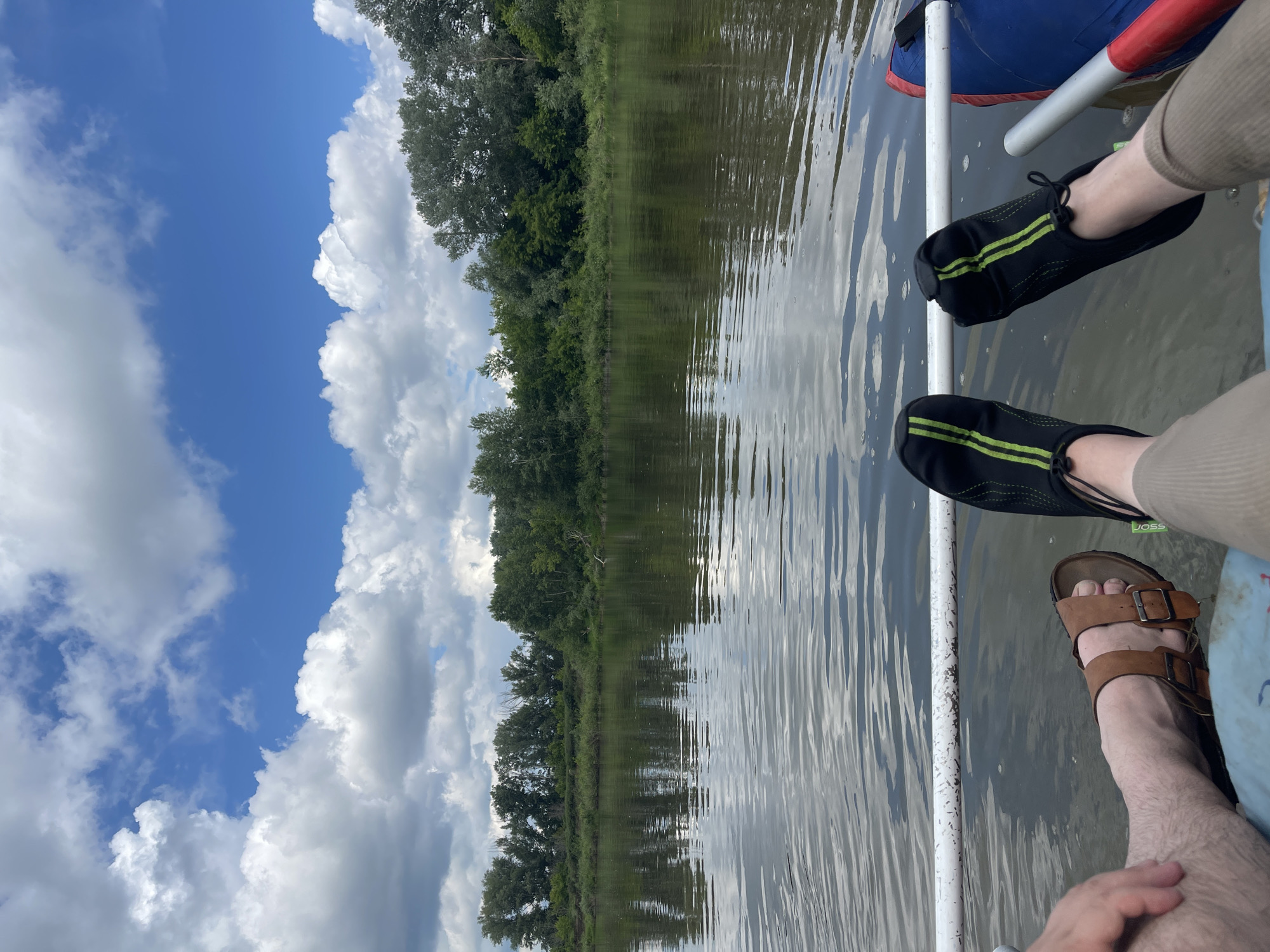 The image depicts a low-angle view of two people's legs and feet from the knees down, sitting in what appears to be a boat on a body of water. The person on the left is wearing brown sandals, while the person on the right is wearing black water shoes with green stripes. Both individuals are positioned on a blue and red inflatable object, likely part of the boat. A white tube or railing runs horizontally across the lower portion of the image.
The background reveals a calm river reflecting a cloudy blue sky and a dense line of green trees and foliage along the far bank, indicating a natural, possibly rural or wilderness setting. The water surface is rippled, suggesting a gentle movement. The scene is set outdoors under daylight, with diffused sunlight filtering through the clouds. The location is identified as the city of Samoradovo, Russia.