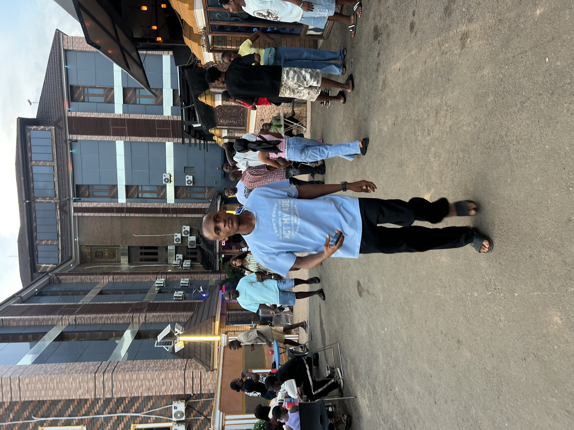 The image depicts an outdoor scene in Obosi, Nigeria, likely a commercial area with multi-story buildings. In the foreground, a young man wearing a light blue t-shirt and black pants walks towards the camera, holding a mobile phone. Behind him, other individuals are visible, some seated at tables, others standing and conversing. The buildings in the background feature brick facades and dark-tinted windows, with visible air conditioning units mounted on their exteriors. The ground is paved with asphalt. The lighting suggests it is either late afternoon or early evening.