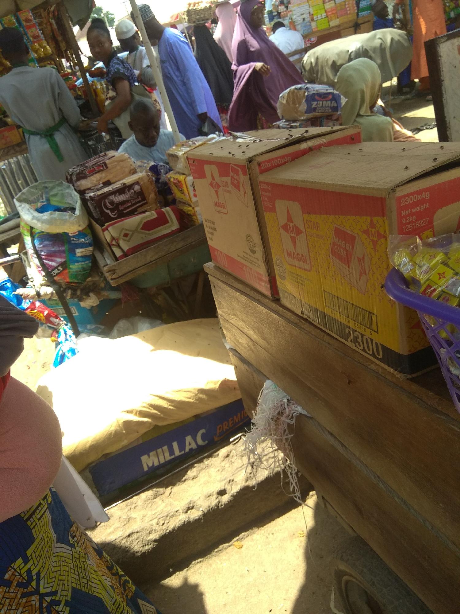 In Jalingo, Nigeria, a street market scene unfolds with a focus on goods displayed for sale. Several people, some in traditional attire, are present. A woman wearing a purple hijab and a flowing purple garment is visible, and another woman in a green hijab is partially obscured. A man in a blue robe stands to the right, and another man in a light blue shirt is seated. The foreground is dominated by stacks of goods, including boxes labeled "MAGGI CUBE" and bags of what appear to be food items. A blue plastic basket filled with small yellow packets is also visible. The market appears to be set up outdoors, with sunlight casting shadows. The ground is sandy, and various improvised structures, possibly temporary stalls, are present. The overall impression is of a bustling marketplace with a variety of products.
