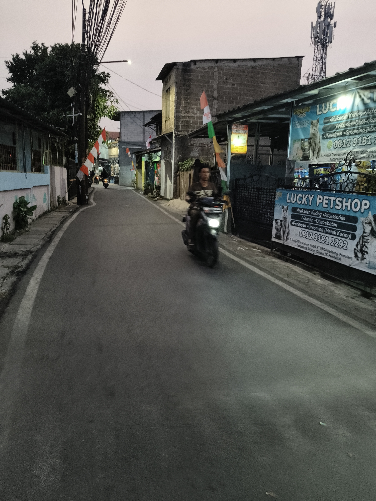 A motorcycle rider is captured in motion on a paved road in South Tangerang, Indonesia, during dusk. The rider is blurred due to movement. To the right of the road, a "LUCKY PETSHOP" banner is visible, advertising pet food, accessories, vitamins, and grooming services. A phone number, "0812 9181 2292", and an address "Jl. Masjid Darussalam No.50 RT 09/04 Kedaung, Pamulang, Tangerang Selatan (Depan TK Pamulang)" are also displayed on the banner. Indonesian flags are seen on poles along the street. The environment appears to be a residential or commercial area with buildings lining the road and a cellular tower visible in the background.