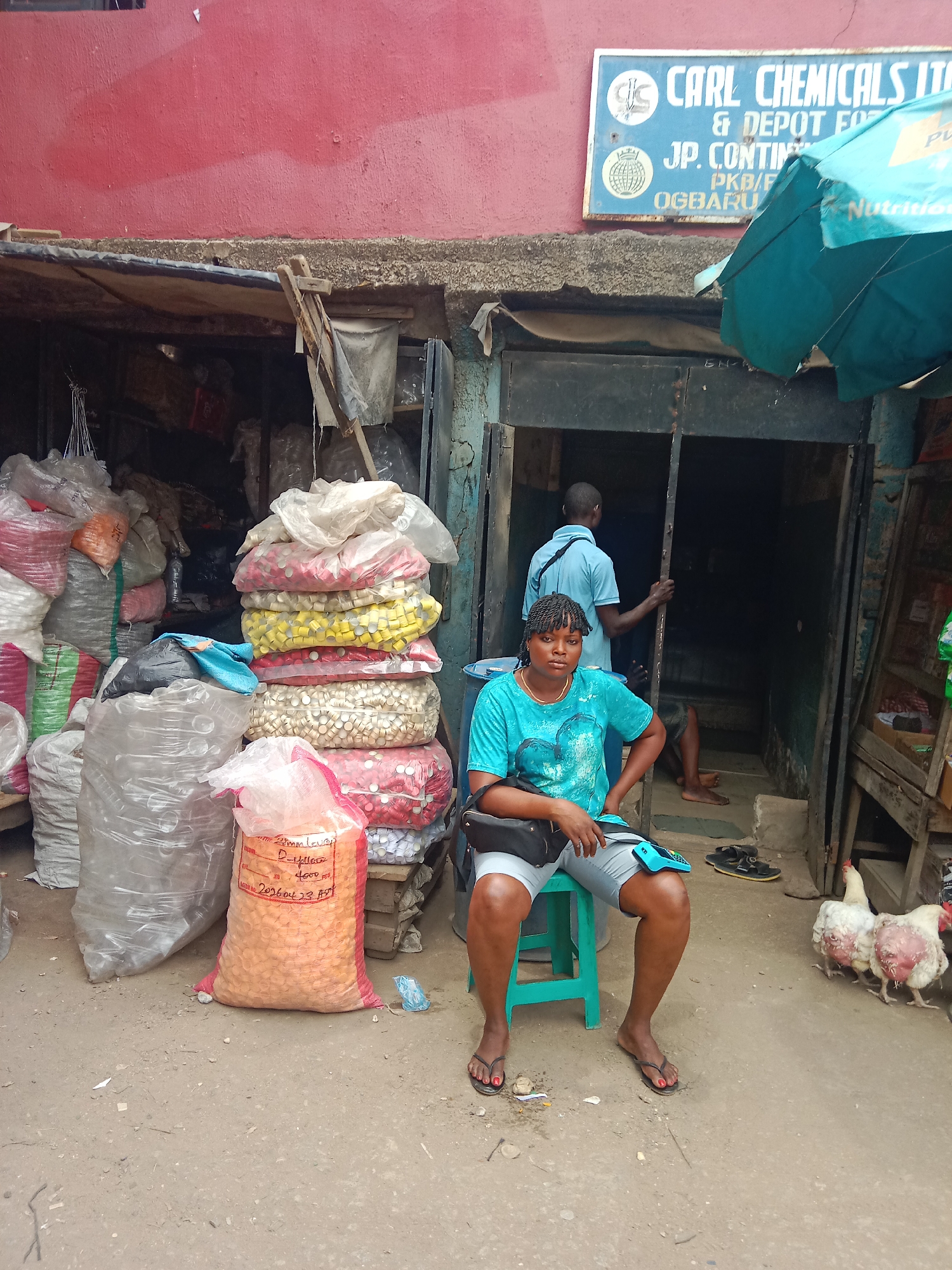 The image shows an outdoor vendor scene in Onitsha, Nigeria. A woman sits on a green stool in front of a building with closed metal doors. The building's exterior wall is a faded red. Above the doors is a sign reading "CARL CHEMICALS IT & DEPOT FO JP. CONTIN PKB/F OGBARU," likely indicating the business name. A man stands inside the doorway behind her. Piles of large, clear plastic bags containing colored plastic bottle caps are stacked to her left. A bag labeled with pricing information is also visible. Two chickens stand to her right. A turquoise umbrella provides shade over part of the scene. The ground is dusty.