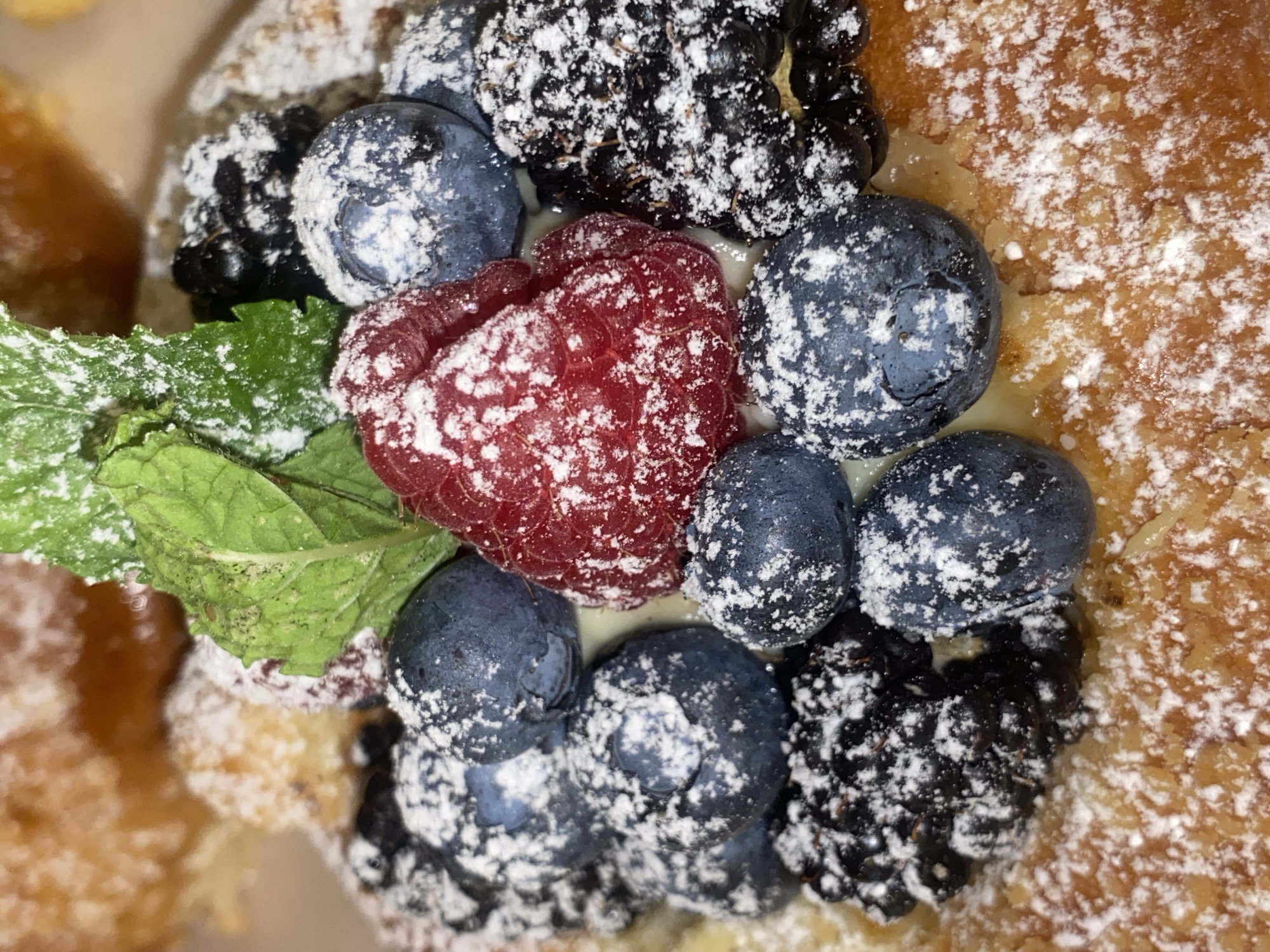 This close-up shot captures a delectable dessert, likely a pastry or tart, generously topped with fresh berries and a dusting of powdered sugar. Prominently featured are bright red raspberries, deep blue blueberries, and dark blackberries, all coated in a fine white powder that accentuates their textures. A few vibrant green mint leaves are interspersed among the berries, adding a fresh contrast. The pastry itself appears golden brown and flaky, suggesting it's freshly baked. The background is out of focus, but hints of more pastry and a creamy filling are visible, creating a shallow depth of field that draws attention to the fruit topping. The lighting is soft and even, highlighting the glistening surfaces of the berries and the delicate powdery coating. There is no visible text or indication of time of day, weather, or specific emotions. The context provided suggests a setting in Warsaw, Poland, implying this could be a treat from a local bakery or cafe.
