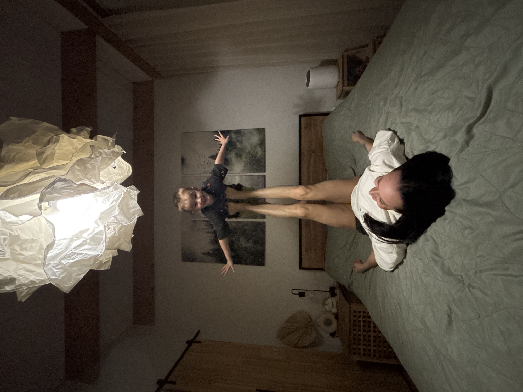 The image shows the interior of a room. A woman with dark hair is lying on a bed covered with a green duvet. Opposite her, in what appears to be a vertical reflection or adjacent image, a child is standing with their arms raised. Between them lies their stretched legs. In the background, a piece of art can be seen hanging on the wall. A lamp with a complex, ruffled shade hangs in the foreground. The room's decor includes wooden accents and a nightstand with decorative items. There is no indication of the location as Chaikovsky, Russia in the visual data.