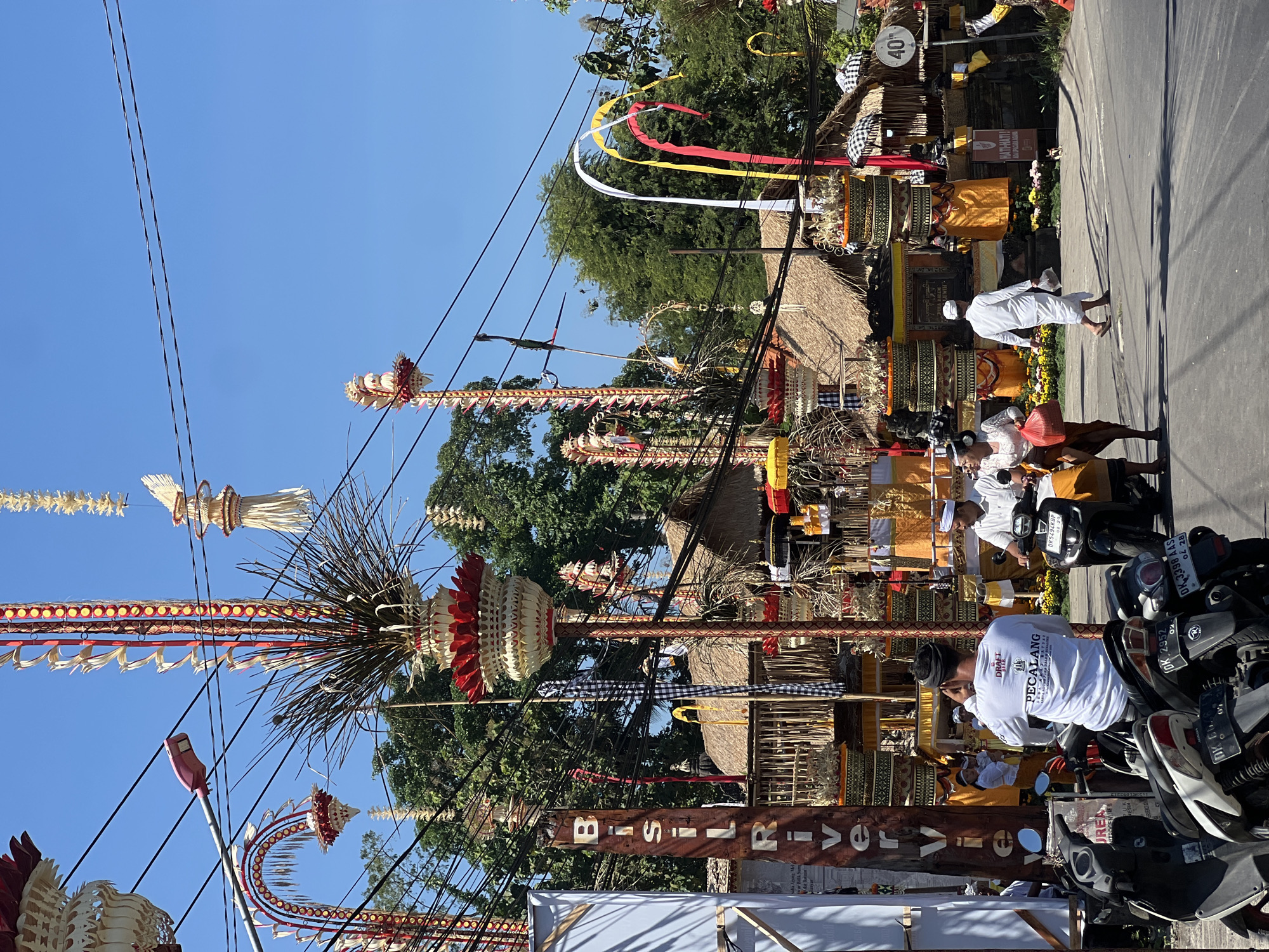 The image captures a street scene in Sukawati, Indonesia, characterized by vibrant decorations and a clear blue sky. Towering, ornate structures, likely religious or ceremonial in nature, are prominent, adorned with intricate woven materials, red and white accents, and what appear to be offerings. These structures are interspersed with numerous electrical wires, some draped low over the street. A tall, narrow wooden sign with white lettering reading "BISIL RIVER" is visible on the left.
Several people are present in the foreground and midground. A man in a white long-sleeved shirt with text on the back, identified by the cropped image as "PECALANG", is seated on a scooter, looking over his shoulder. To his right, a man and a woman on a scooter are moving away from the camera. The woman is wearing a white top and an orange skirt, and the man is wearing a white shirt and a white head covering. Further back, a man in a white outfit, possibly a traditional sarong and shirt, walks across the street carrying a plastic bag. On the right side of the street, another person in a similar white outfit is partially visible. Several motorcycles are parked in the foreground. The background features traditional Balinese-style buildings with thatched roofs and brightly colored draped fabrics, suggesting a festive occasion or market area. Flowers line the front of some structures. A speed limit sign indicating "40" is mounted on a pole on the right side of the street.
