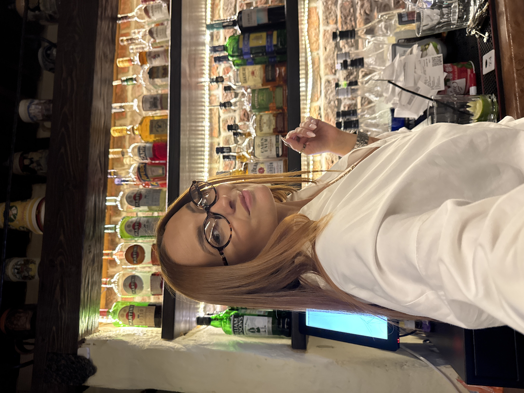 Here is a description of the image:
The image shows a woman with reddish-brown hair wearing glasses and a white shirt. She appears to be behind a bar. In the background, there are shelves filled with various liquor bottles. There is also a computer monitor near the lower part of the image. The setting suggests a bar or restaurant environment. It is not possible to identify if the location is Chaikovsky, Russia from the picture alone.
