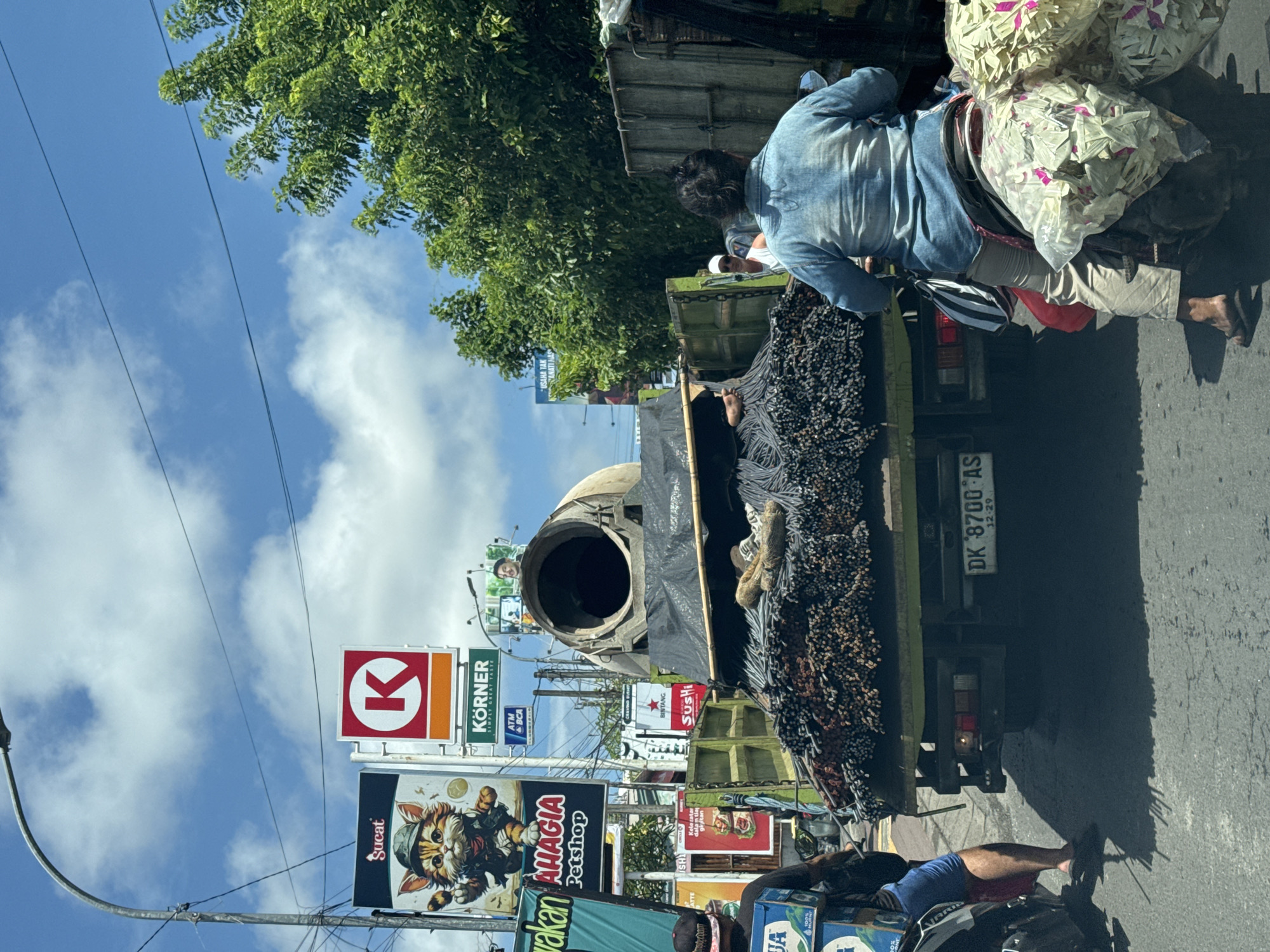 The image depicts a street scene in Kuta Selatan, Indonesia, showcasing a truck heavily laden with metal rods and an industrial cylindrical object, possibly part of construction equipment, in its truck bed. A person in a light blue shirt stands near the truck's load. A second person is seated on a motorcycle behind the truck, carrying two large bags. The background is filled with billboards advertising various products and services, including "Sucat," "AHAGIA Petshop," "KÖRNER," "BINTANG," and "Sushi." A license plate is visible on the rear of the truck. The sky is partly cloudy.