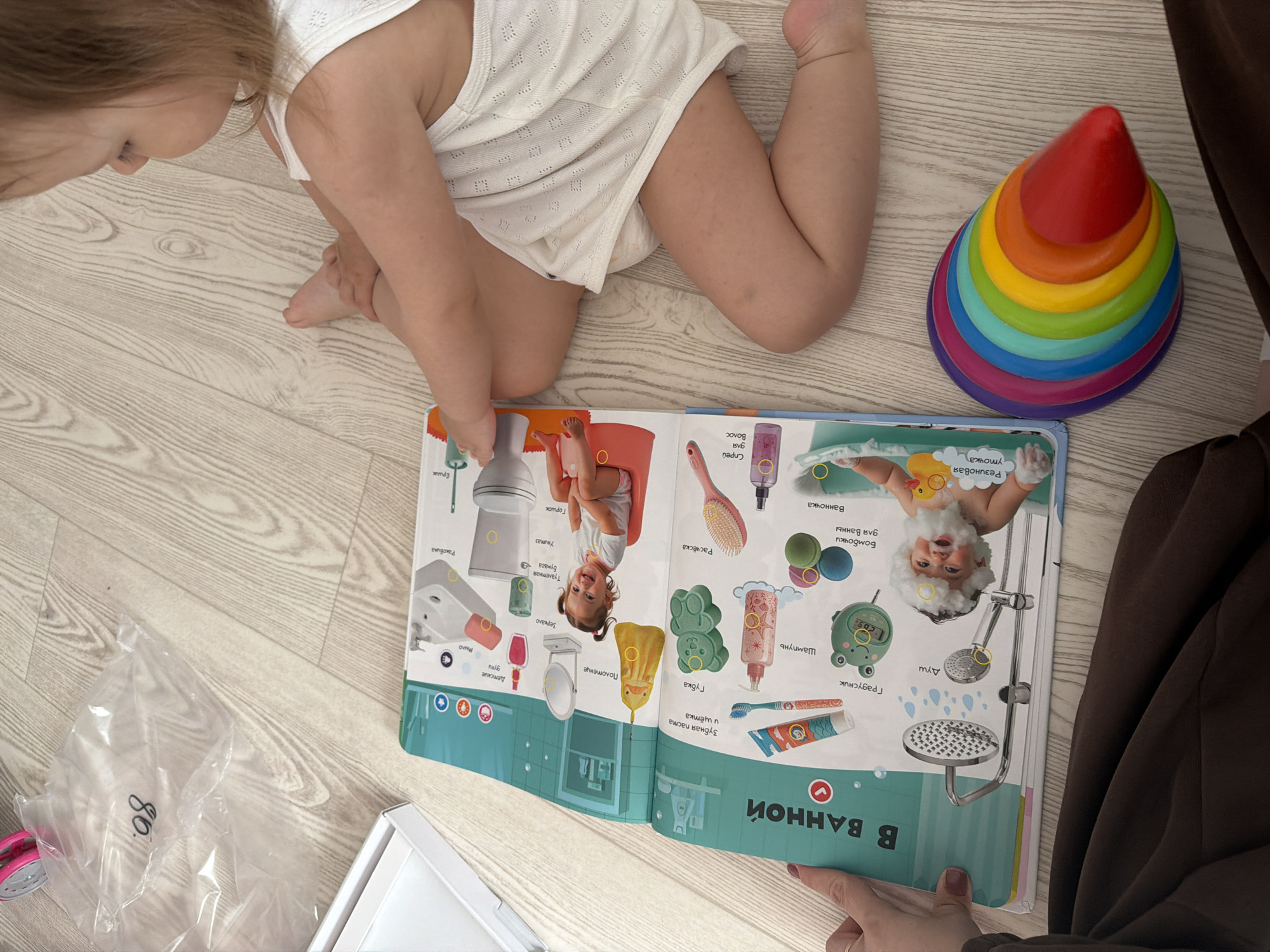 A child in a white outfit is sitting on the floor, looking at an open book titled "В ВАННОЙ" (In the Bathroom), which contains illustrations of bathroom items. A colorful stacking toy is next to the child. A brown curtain is visible in the background. A plastic bag and a white box are in the lower-left corner. The photo was taken indoors.
