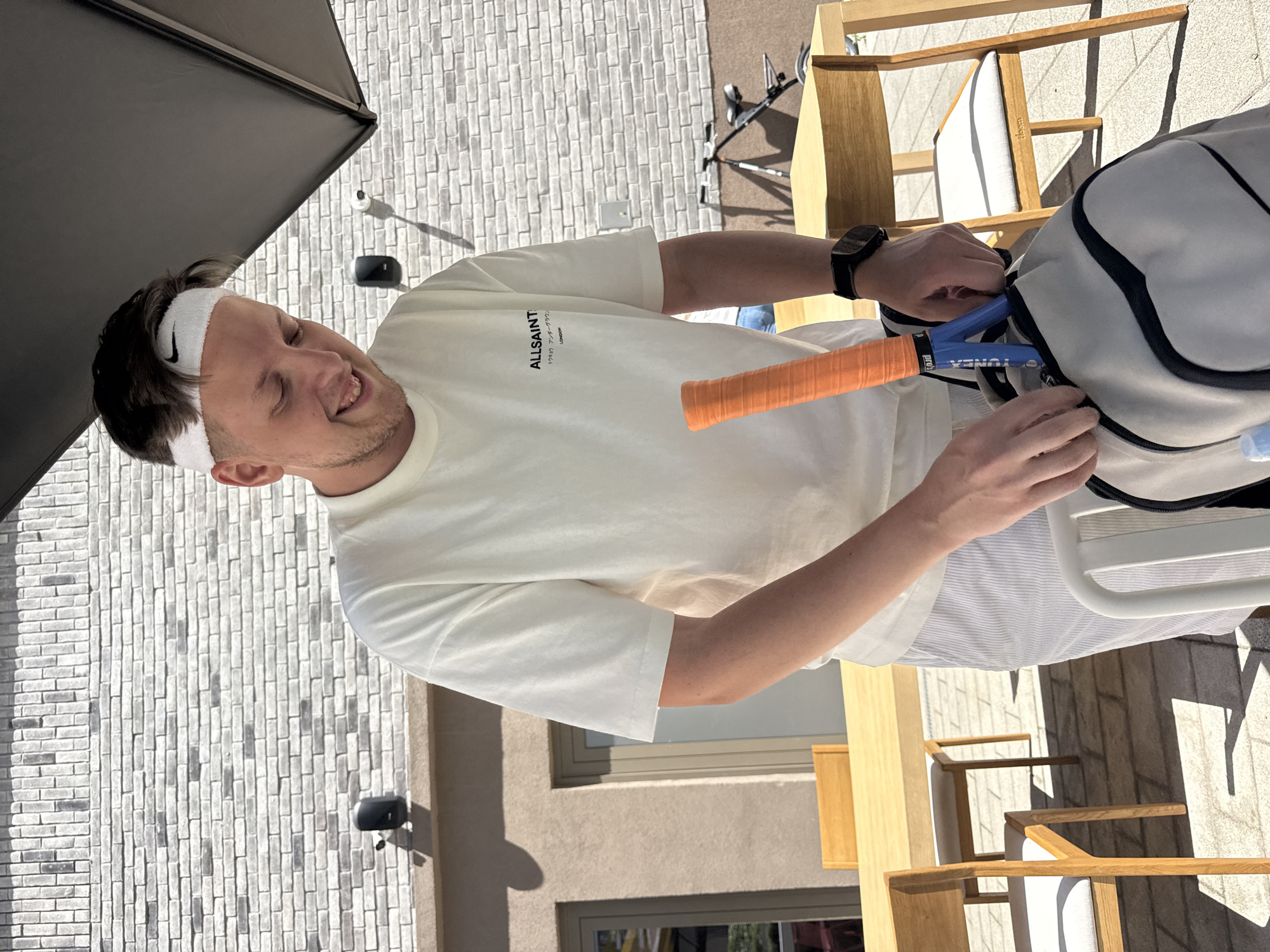 A fair-skinned man with a white headband is smiling while putting a blue Yonex tennis racket with an orange grip into a white tennis bag. The man is wearing a white t-shirt and is outside on a patio with gray brick walls and a tan-colored building in the background. The scene includes outdoor furniture and is presumably in the city of Belgrade, Serbia.